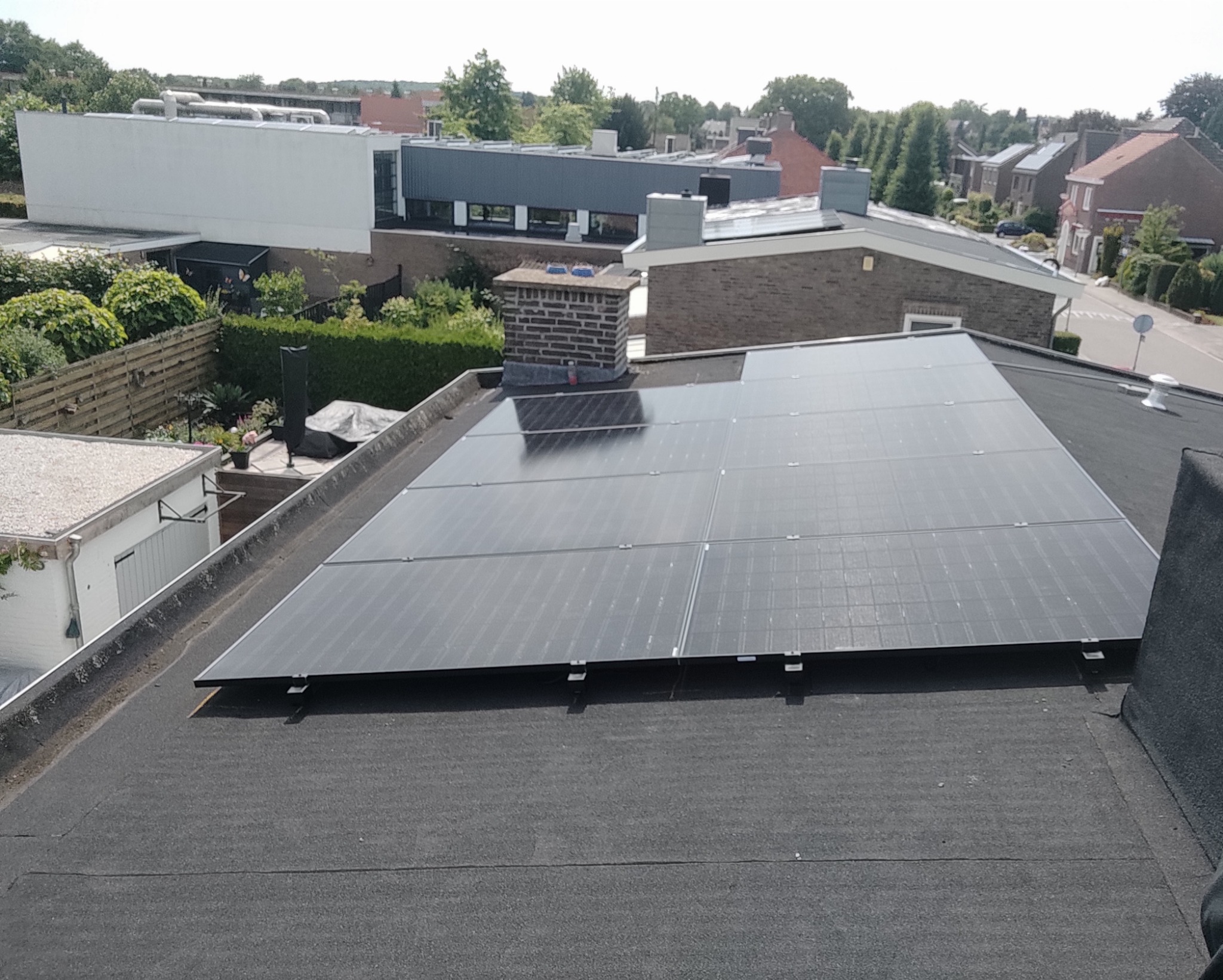 Panele słoneczne na płaskim dachu budynku mieszkalnego, widok z góry, w tle budynki i zieleń.