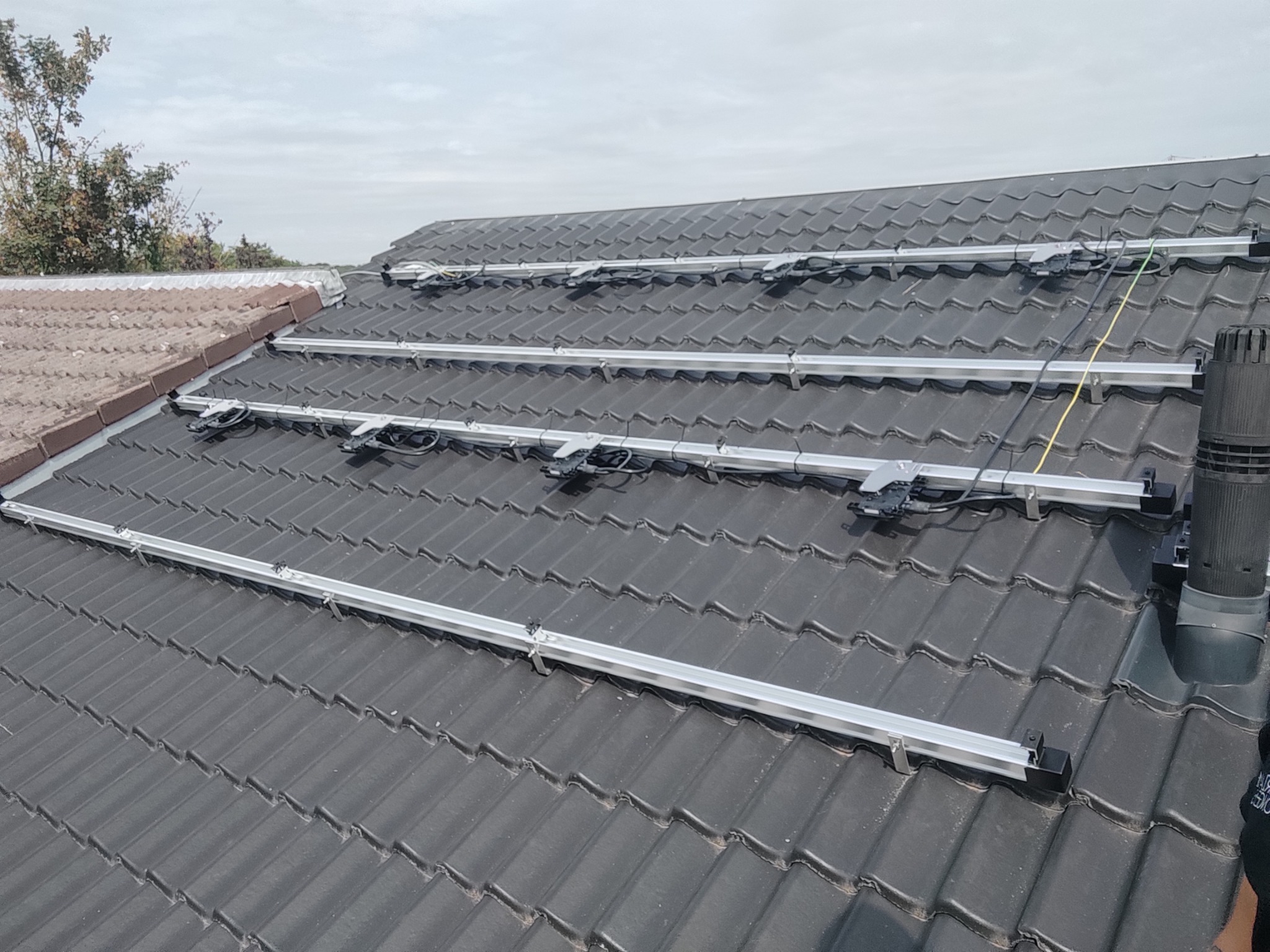 Montaż stelażu pod panele na dachu z ciemnej dachówki ceramicznej, widoczne kable i komin wentylacyjny.