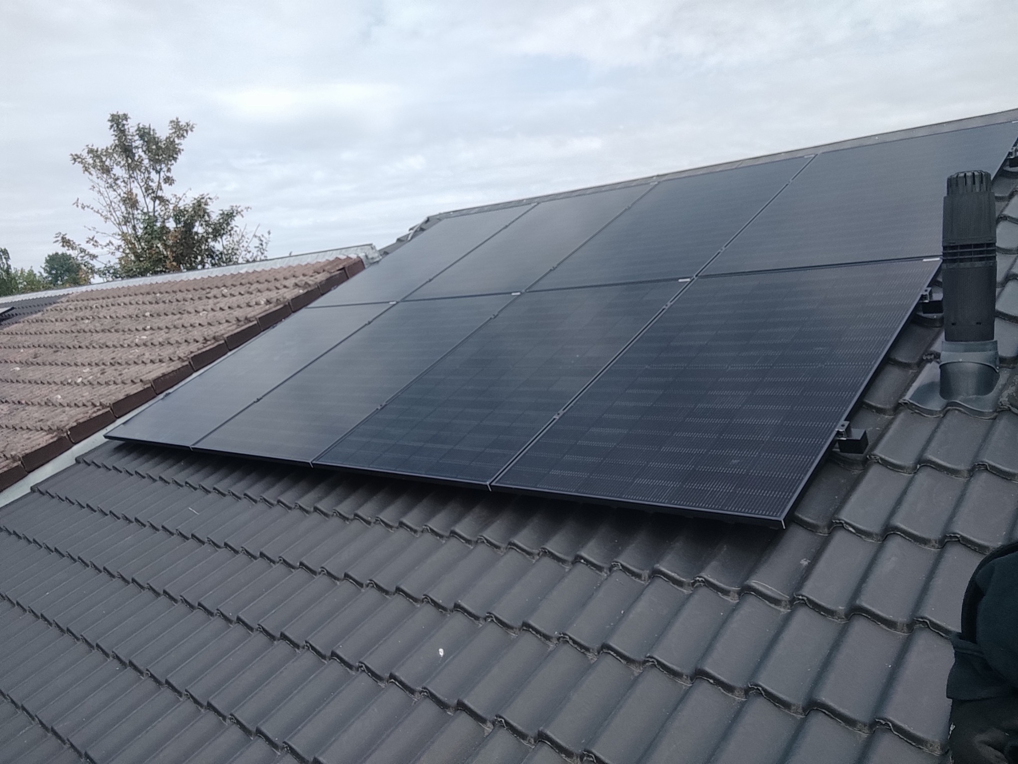 Czarny panel na dachu z ciemnej dachówki, widoczny komin wentylacyjny i fragment dachu z jasnej dachówki w tle, pochmurne niebo.