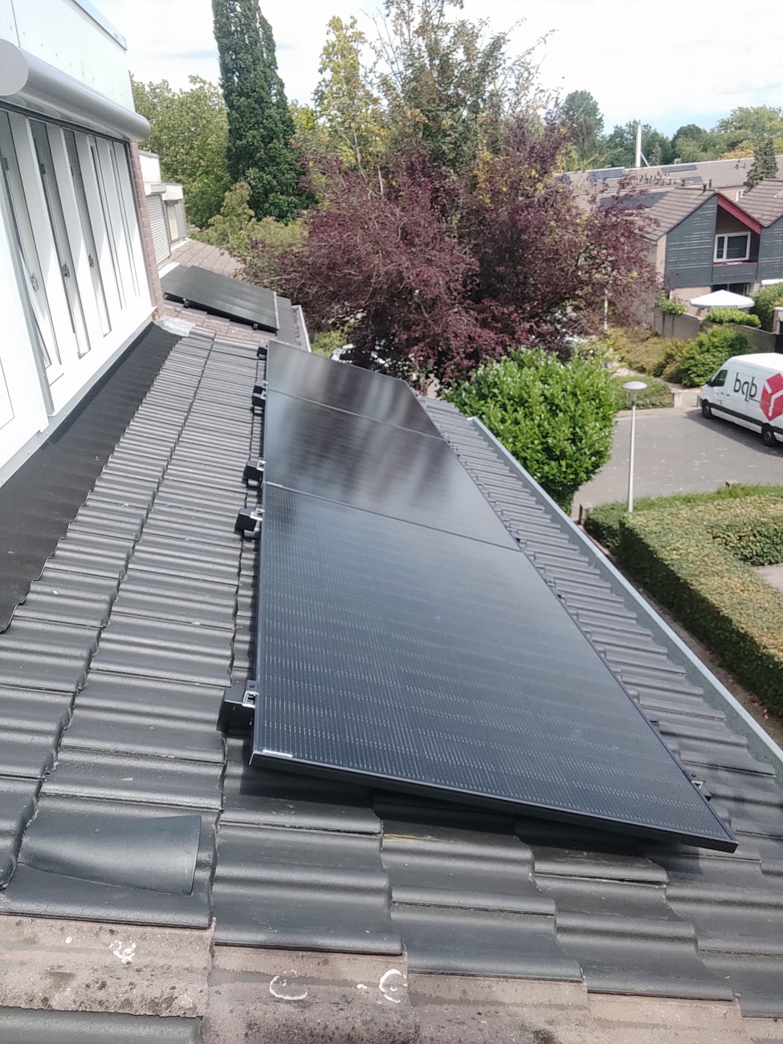 Czarne panele fotowoltaiczne zamontowane na szarym, ceramicznym dachu domu jednorodzinnego, widok z góry, w tle zieleń drzew i zabudowa mieszkalna.