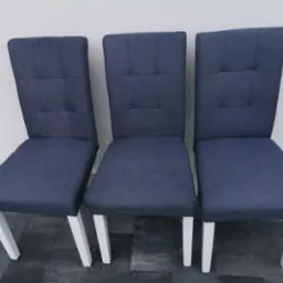 Trzy identyczne krzesła tapicerowane ciemnoniebieską tkaniną, z białymi, ukośnymi nogami, ustawione obok siebie na szarym bruku przed białą ścianą.