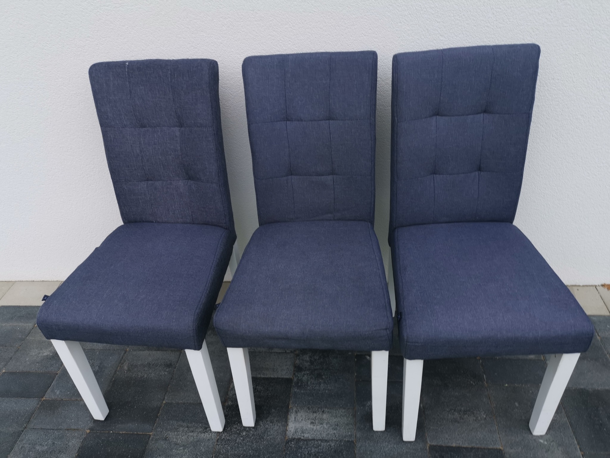 Trzy identyczne krzesła tapicerowane ciemnoniebieską tkaniną, z białymi, ukośnymi nogami, ustawione obok siebie na szarym bruku przed białą ścianą.
