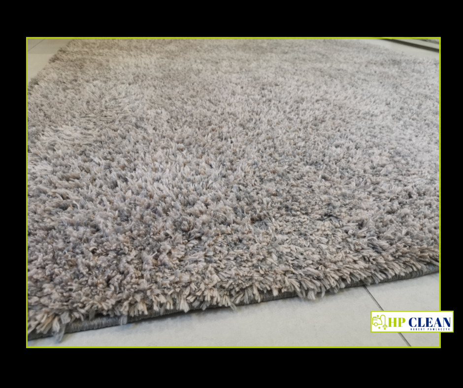 Szary, puszysty dywan z długim włosiem, widoczny z bliska, z logo firmy sprzątającej w prawym dolnym rogu.