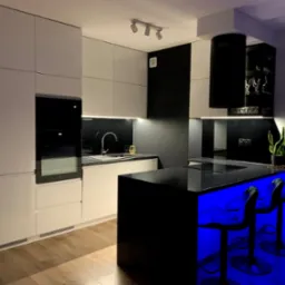 Nowoczesna kuchnia z białymi szafkami, czarnym blatem i wyspą podświetloną niebieskim światłem, drewniana podłoga, minimalistyczny design.