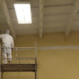 FastPainting - Pracownik w kombinezonie ochronnym na rusztowaniu maluje ścianę na żółto, widoczny fragment pomalowanego sufitu z oświetleniem.