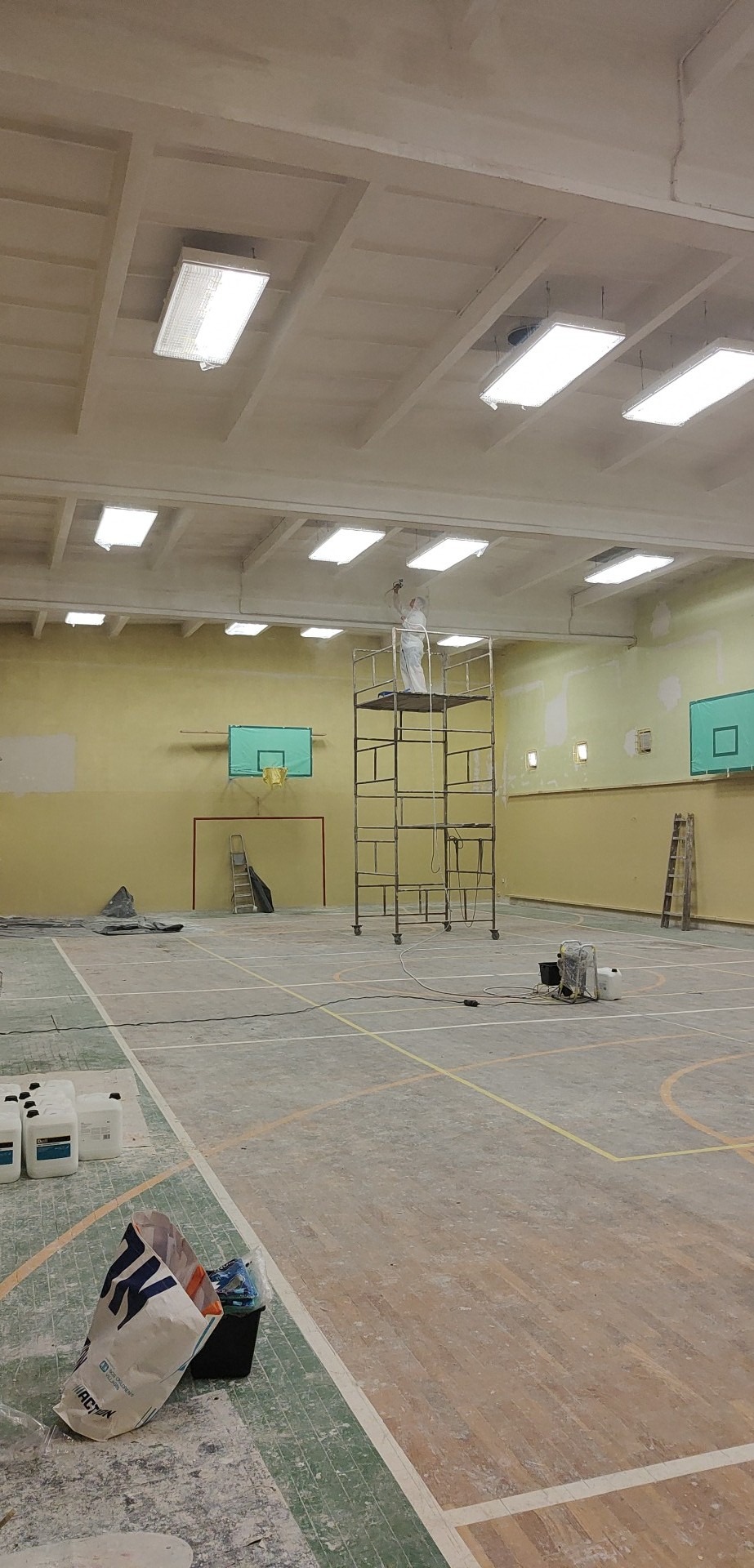 Pracownik w kombinezonie ochronnym maluje sufit hali sportowej z podnośnika, widoczne ślady farby na podłodze, sprzęt malarski, kosze do koszykówki i oświetlenie sufitowe.