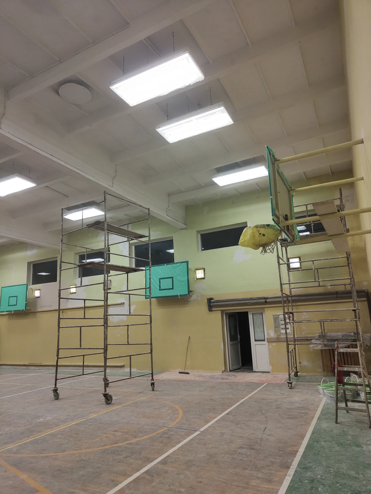 Wnętrze sali gimnastycznej z rusztowaniami podczas prac wykończeniowych, widoczne ślady szpachlowania na ścianach i suficie, zabezpieczone kosze do koszykówki.