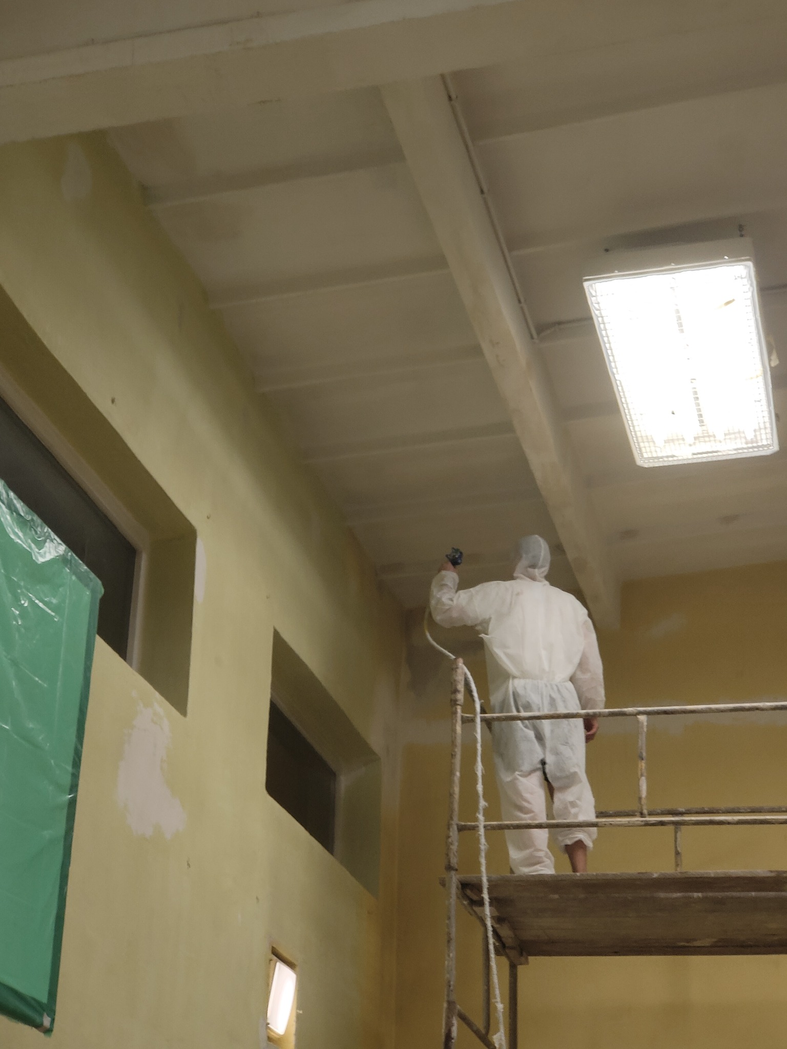 Pracownik w kombinezonie ochronnym na rusztowaniu maluje sufit w pomieszczeniu za pomocą pistoletu natryskowego. Widoczne fragmenty ściany po szpachlowaniu.