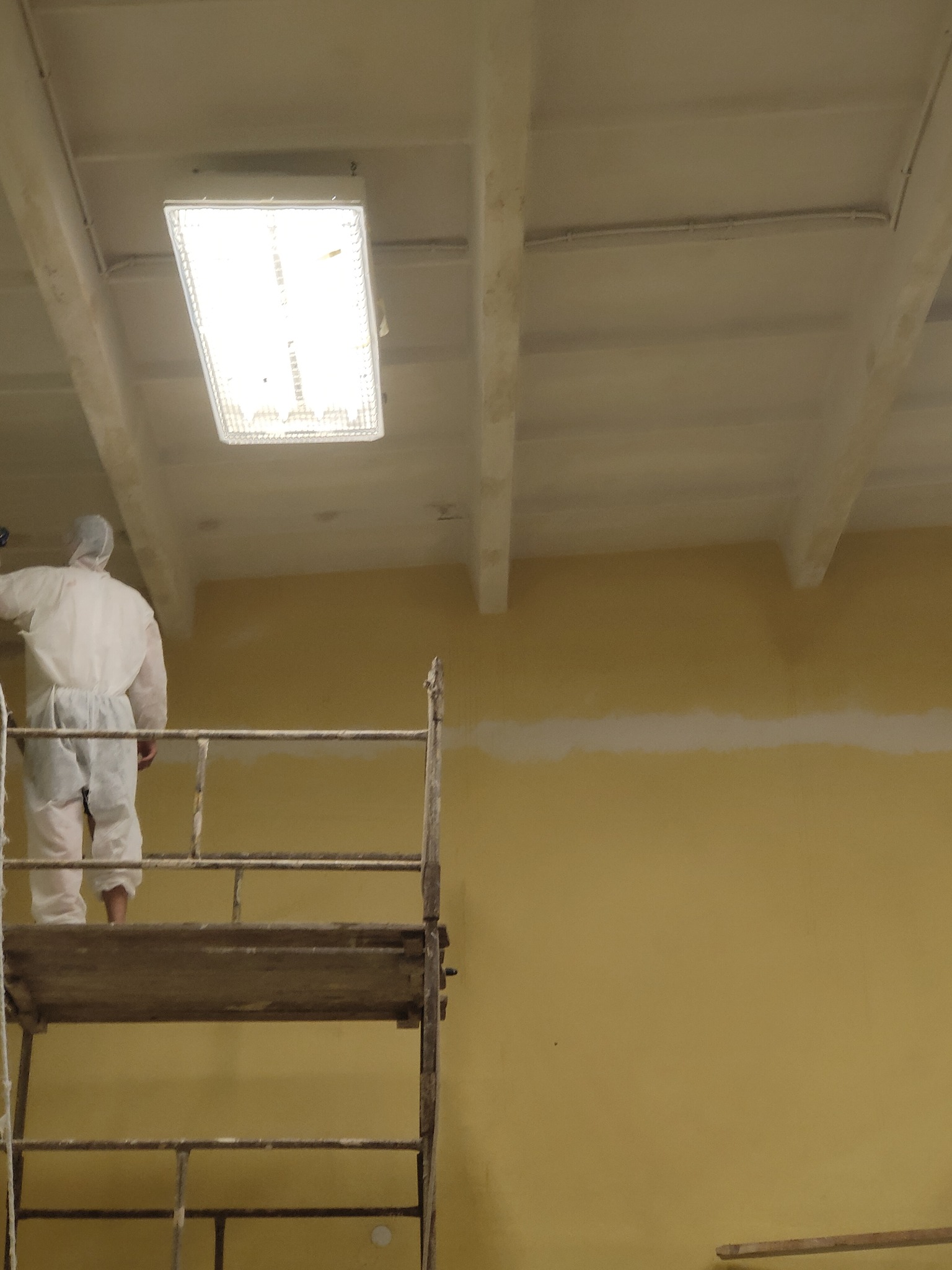 Pracownik w kombinezonie ochronnym na rusztowaniu maluje ścianę na żółto, widoczny fragment pomalowanego sufitu z oświetleniem.
