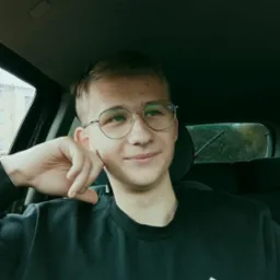 Portret młodego mężczyzny w okularach, siedzącego w samochodzie w pochmurny dzień, z kroplami deszczu na szybie.