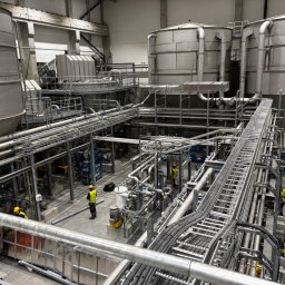 GÓRAL REALIZACJE SPÓŁKA Z OGRANICZONĄ ODPOWIEDZIALNOŚCIĄ - Wnętrze hali przemysłowej z rozbudowaną instalacją rurociągów i zbiorników, pracownicy w żółtych kaskach, widok z góry na konstrukcję.