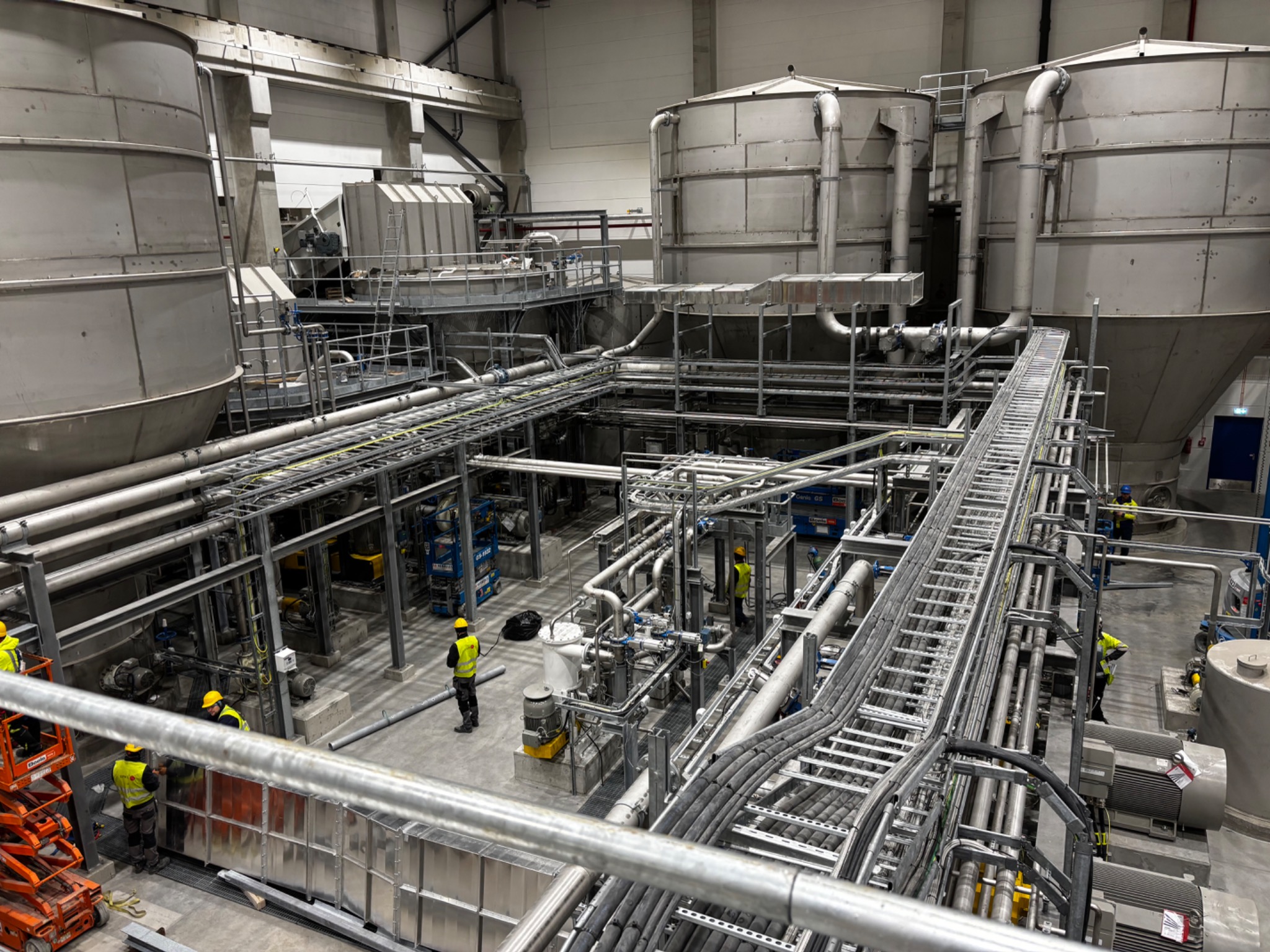 Wnętrze hali przemysłowej z rozbudowaną instalacją rurociągów i zbiorników, pracownicy w żółtych kaskach, widok z góry na konstrukcję.