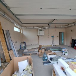 Piotr Skoneczny HydraWro Installation - Wnętrze w trakcie montażu instalacji wentylacyjnej z widocznymi rurami wentylacyjnymi pod sufitem, centralą wentylacyjną WANAS, materiałami budowlanymi i narzędziami na podłodze.