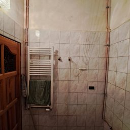 Piotr Skoneczny HydraWro Installation - Biały grzejnik łazienkowy drabinkowy z ręcznikiem na tle kafelkowanej ściany i miedzianych rur instalacji grzewczej w łazience.