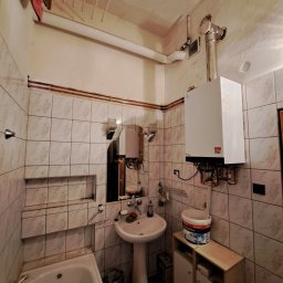 Piotr Skoneczny HydraWro Installation - Łazienka z zamontowanym na ścianie, białym piecem grzewczym, rurami miedzianymi, umywalką na postumencie, wanną i wiadrem farby na szafce.