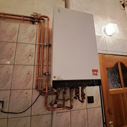 Piotr Skoneczny HydraWro Installation - Biały piec grzewczy Viessmann zamontowany na ścianie z marmurkowymi płytkami, widoczne miedziane rury instalacji grzewczej i fragment drewnianych drzwi.