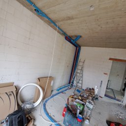 Piotr Skoneczny HydraWro Installation - Wnętrze w trakcie instalacji hydraulicznej: niebieskie rury rozprowadzone po suficie i ścianach surowego budynku, drabina oparta o ścianę, narzędzia i materiały budowlane rozłożone na podłodze.