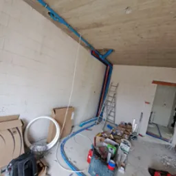 Wnętrze w trakcie instalacji hydraulicznej: niebieskie rury rozprowadzone po suficie i ścianach surowego budynku, drabina oparta o ścianę, narzędzia i materiały budowlane rozłożone na podłodze.
