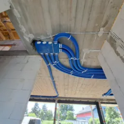 Niebieskie rury wentylacyjne pod sufitem w surowym stanie, widoczne elementy konstrukcyjne budynku w trakcie budowy, białe przewody elektryczne i puszka rozdzielcza.