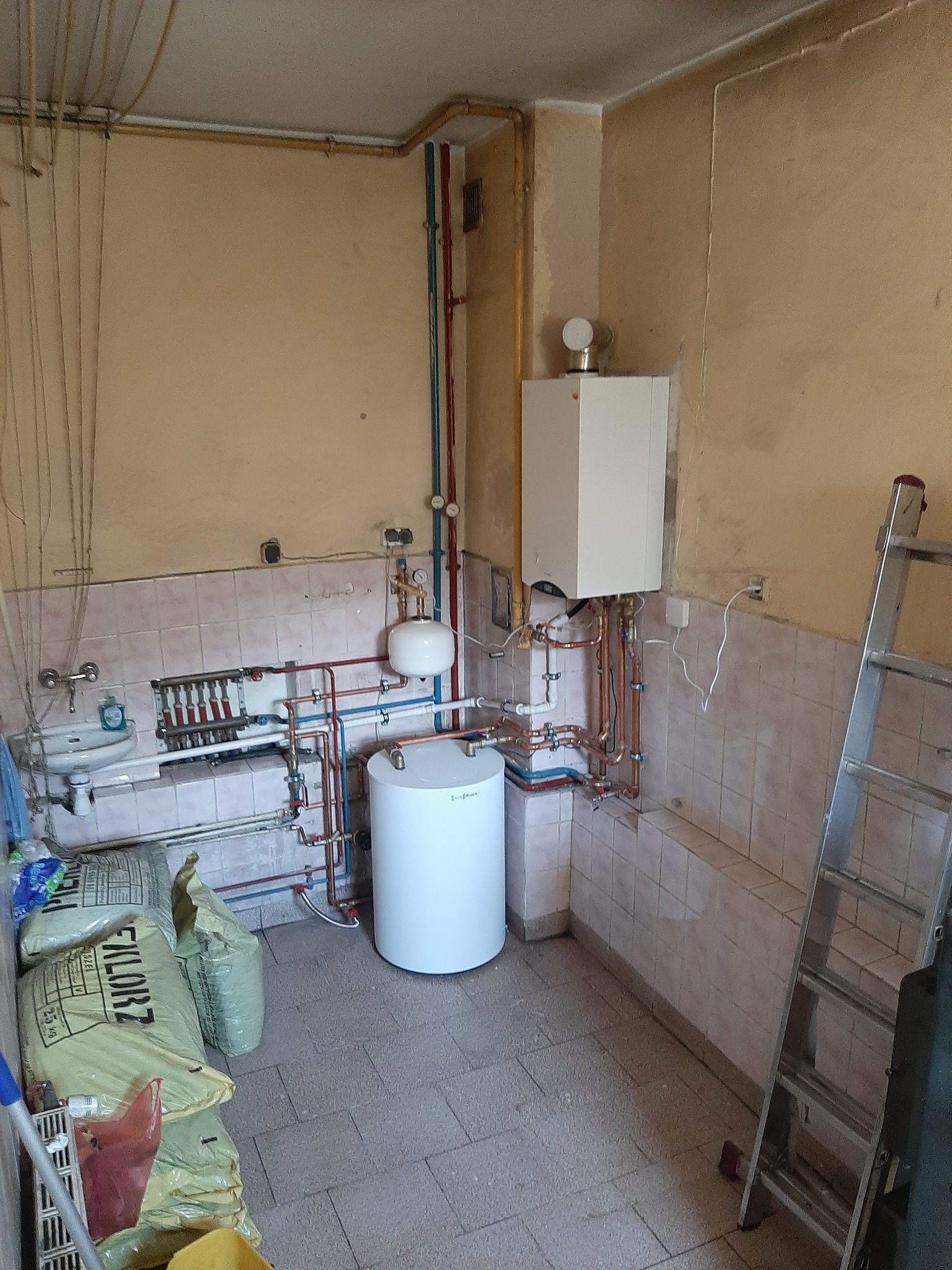 Kotłownia w trakcie instalacji: biały zasobnik ciepłej wody, piec gazowy, rozdzielacz podłogowy, rury miedziane i drabina monterska, worki z zaprawą budowlaną w tle.