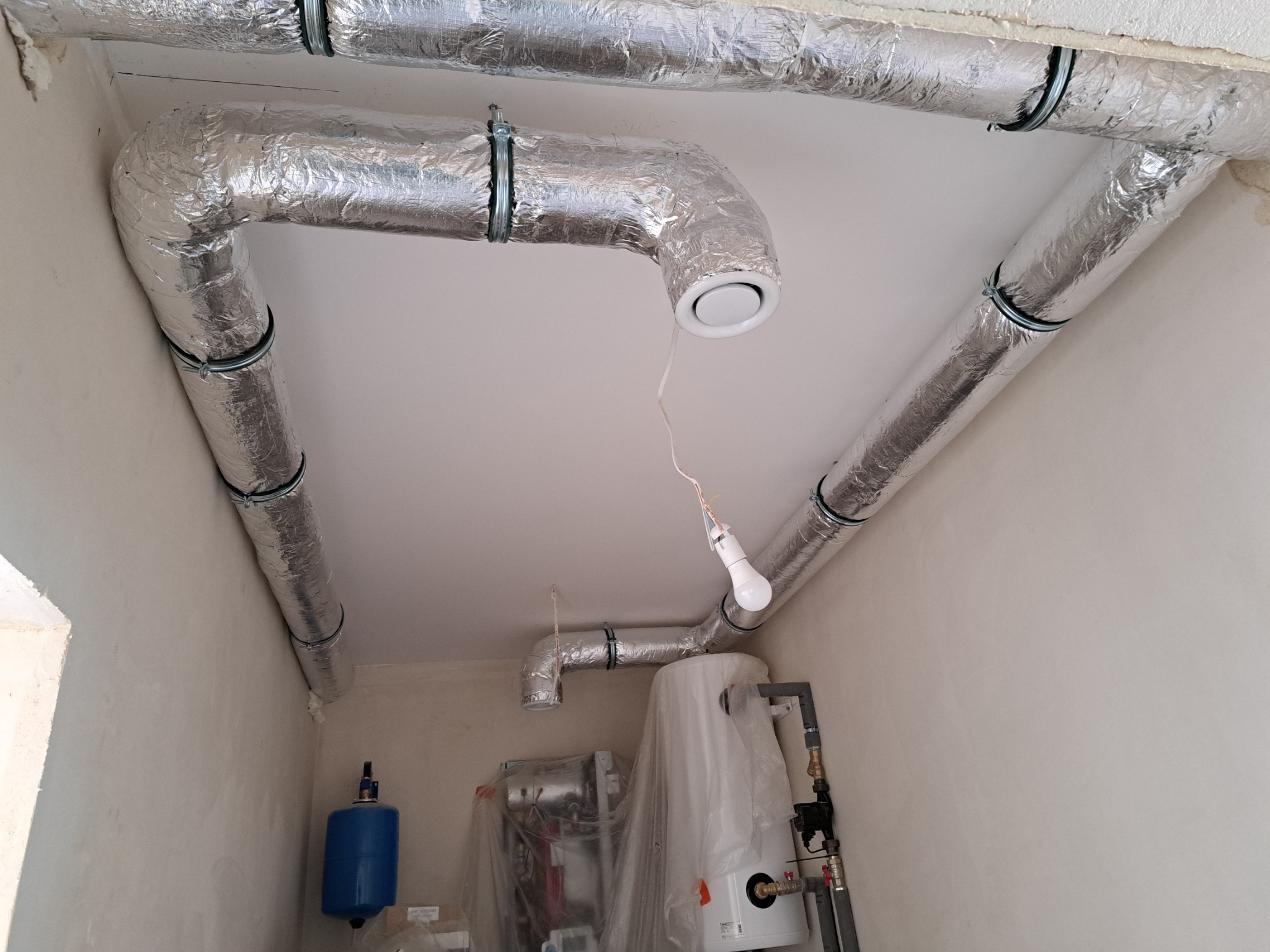 Widok z dołu na izolowane, aluminiową folią przewody wentylacyjne pod sufitem, zakończone okrągłym nawiewnikiem, oraz stojący obok zbiornik na wodę osłonięty folią ochronną.
