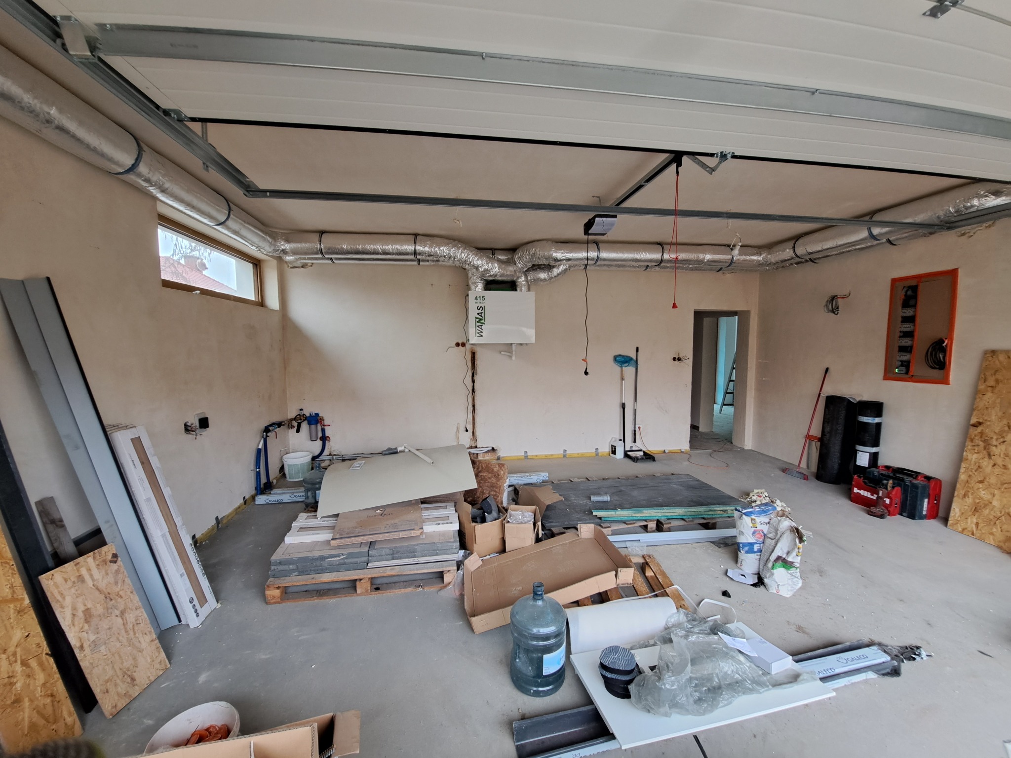 Wnętrze garażu w trakcie instalacji systemu wentylacji mechanicznej z rekuperacją, widoczne kanały wentylacyjne pod sufitem, rekuperator na ścianie, materiały budowlane i narzędzia w nieładzie.