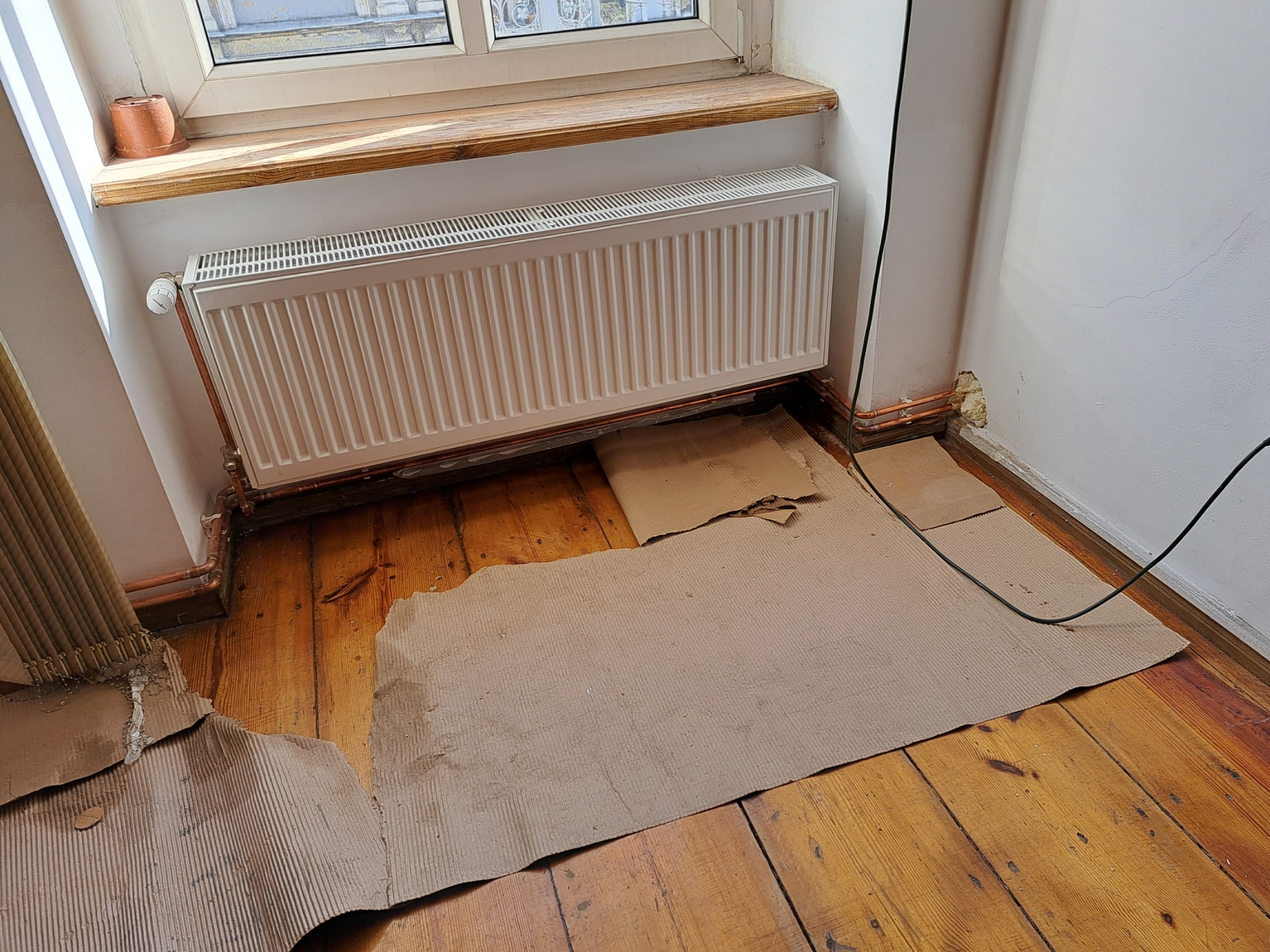 Biały grzejnik panelowy zamontowany pod oknem, zabezpieczony kartonem na drewnianej podłodze podczas prac remontowych, widoczne miedziane rury instalacji grzewczej.