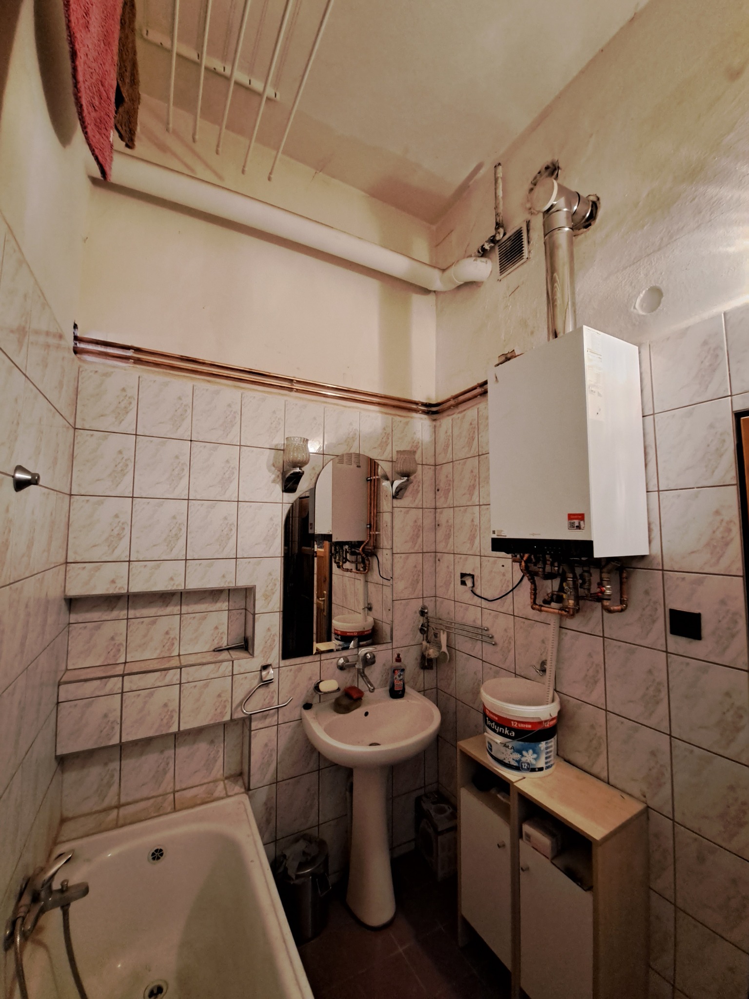 Łazienka z zamontowanym na ścianie, białym piecem grzewczym, rurami miedzianymi, umywalką na postumencie, wanną i wiadrem farby na szafce.