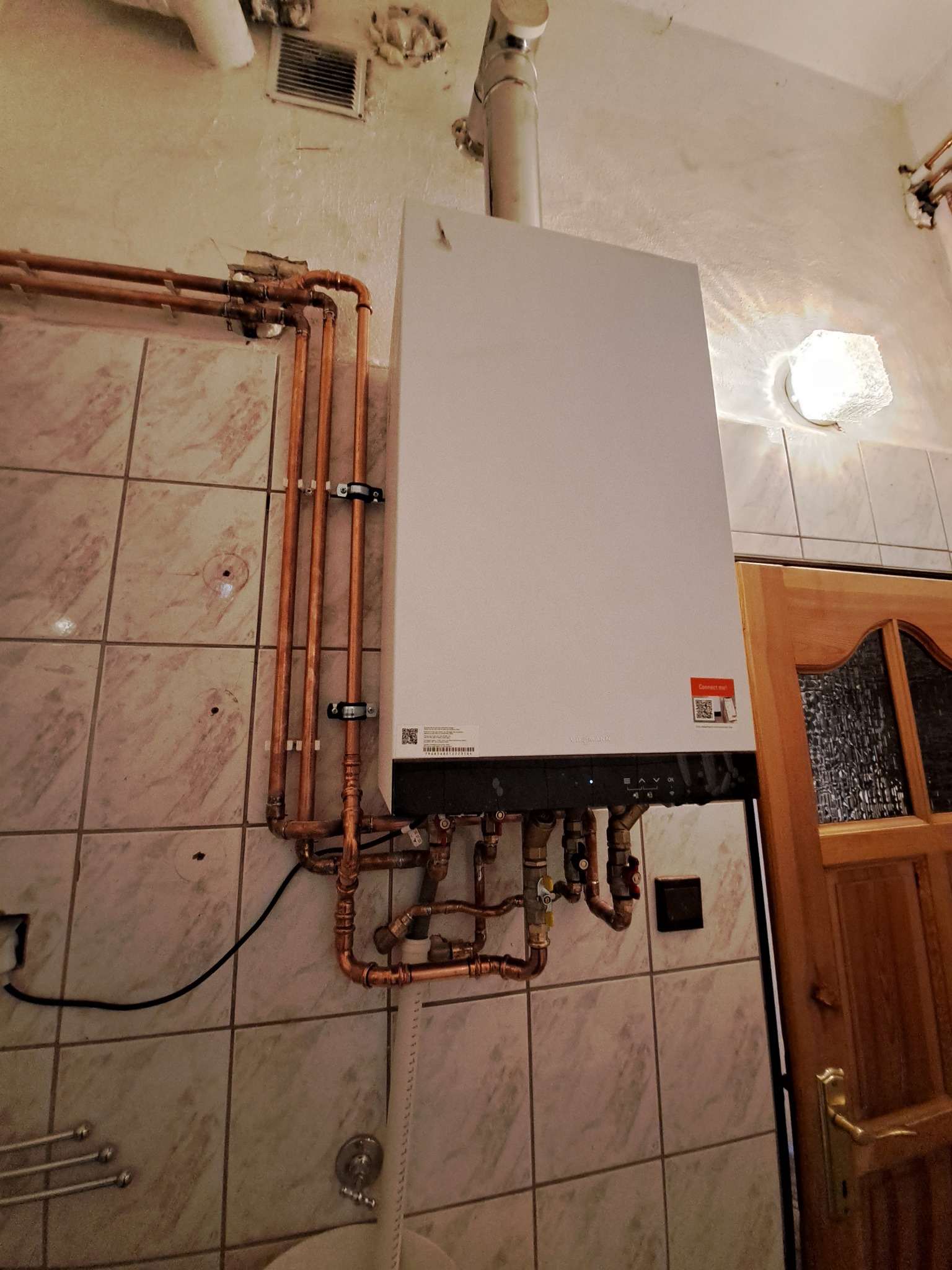 Biały piec grzewczy Viessmann zamontowany na ścianie z marmurkowymi płytkami, widoczne miedziane rury instalacji grzewczej i fragment drewnianych drzwi.