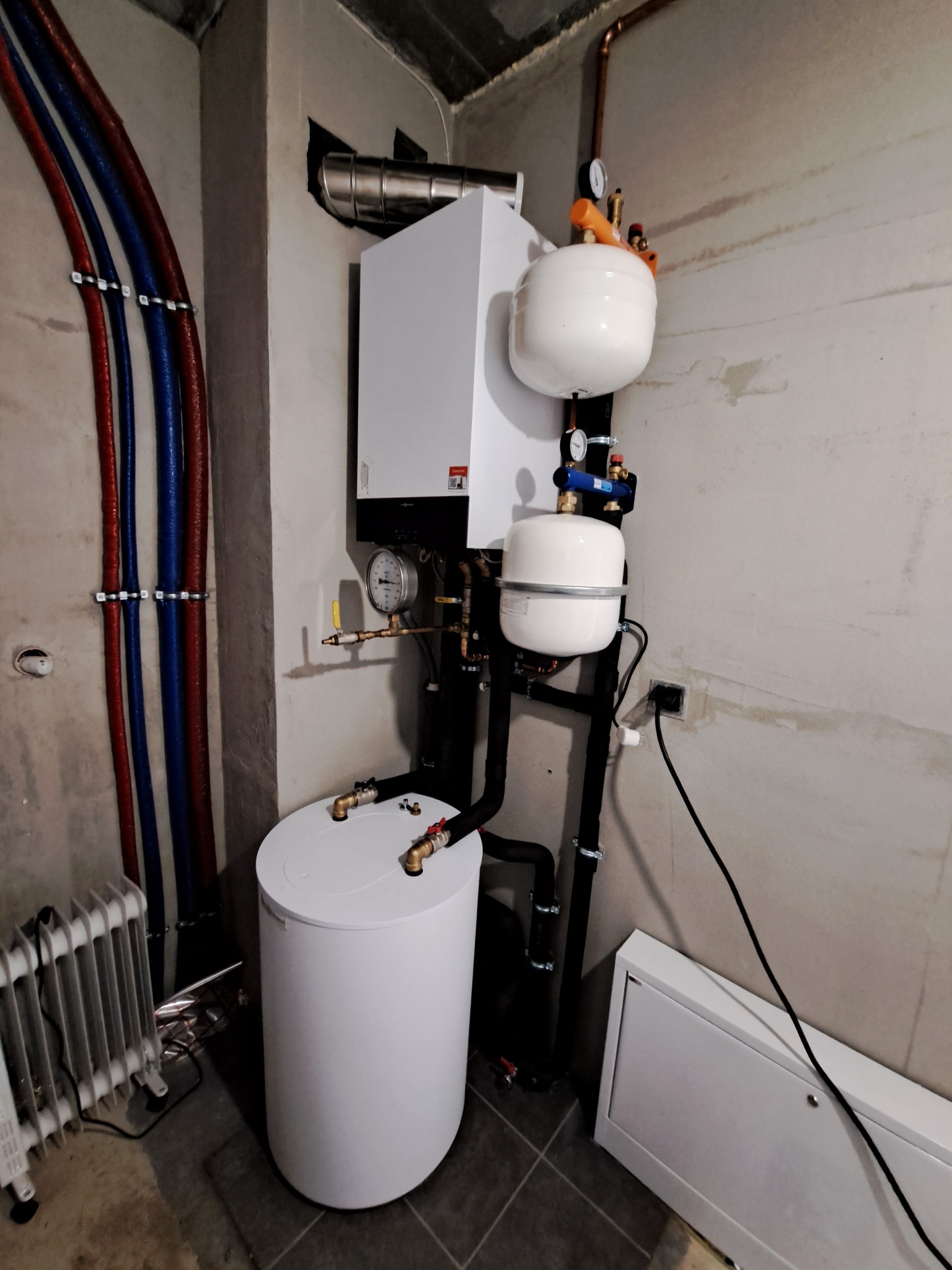Biały piec grzewczy z zbiornikiem buforowym i naczyniami przeponowymi w surowym pomieszczeniu z widocznymi rurami instalacji CO i grzejnikiem żeberkowym.