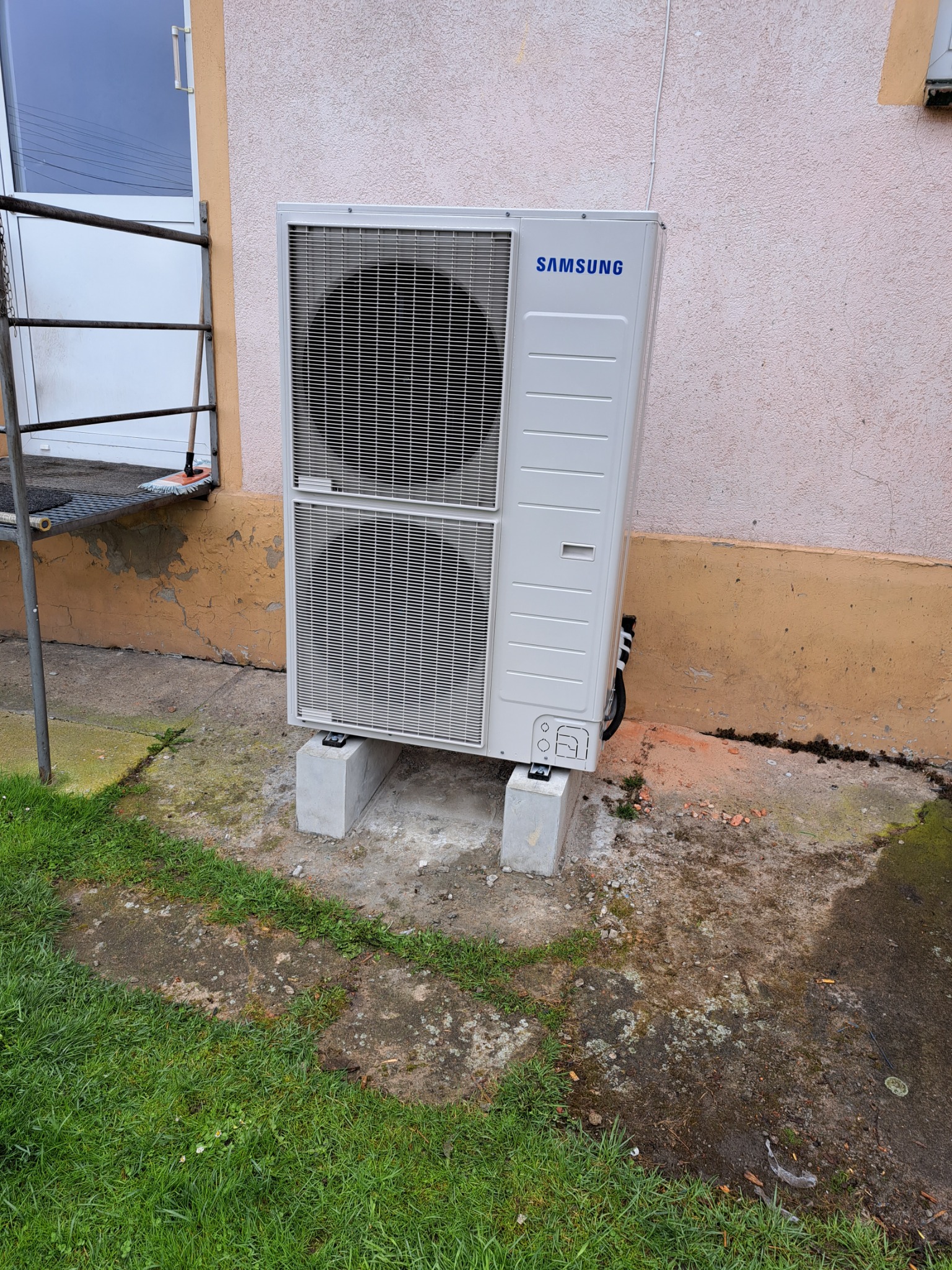 Biała, zewnętrzna jednostka klimatyzacji marki Samsung, umieszczona na betonowych blokach przy ścianie budynku z widocznymi śladami wilgoci i zielenią w tle.