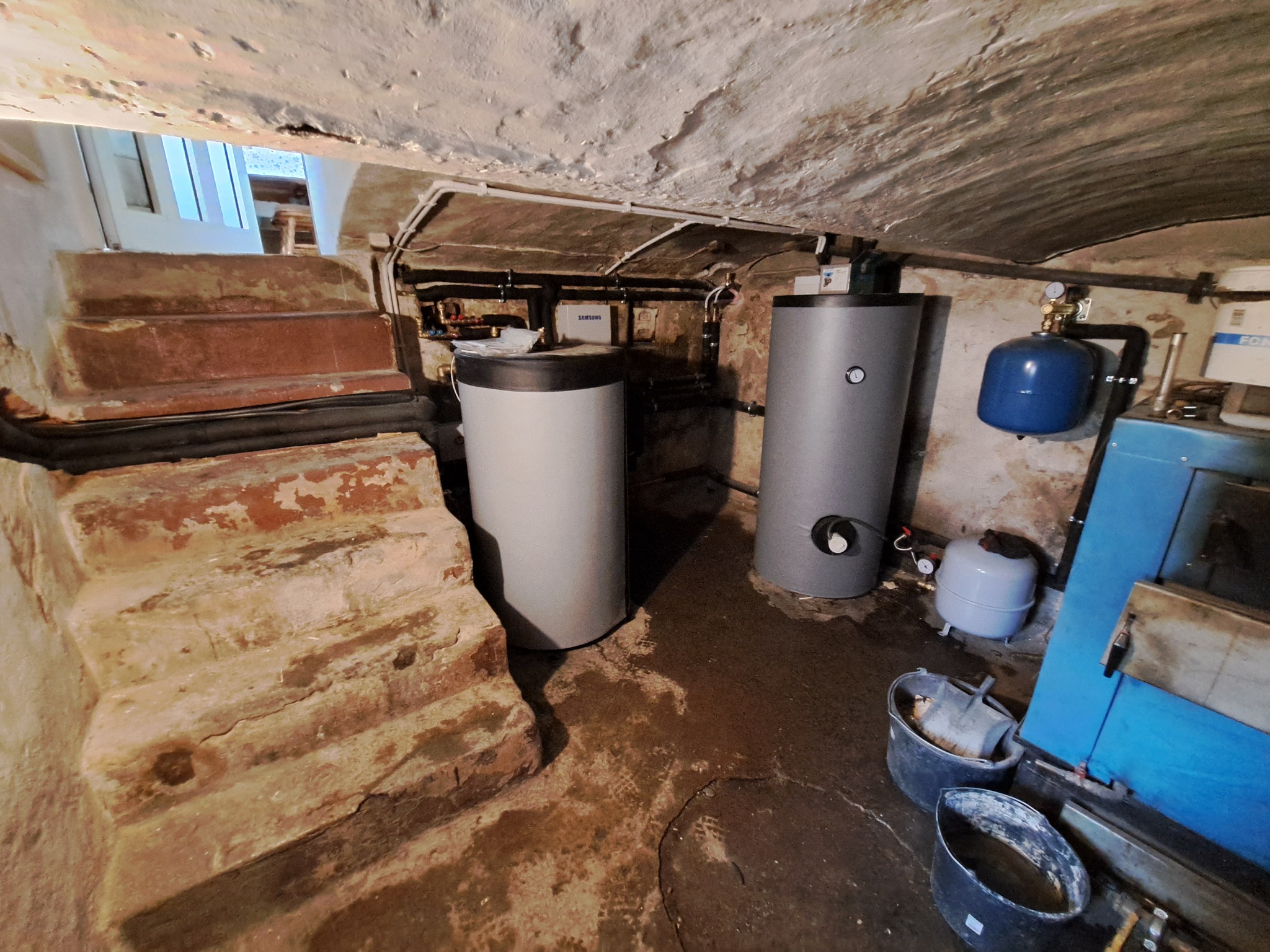 Dwa cylindryczne zbiorniki w piwnicy z ceglanymi schodami, rurami na suficie i dodatkowym sprzętem hydraulicznym, w tym niebieski zbiornik ciśnieniowy i wiaderka.