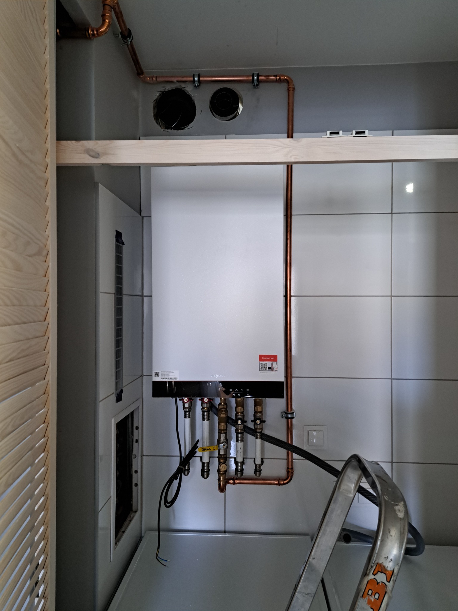 Biały piec grzewczy Viessmann zamontowany na ścianie z białych płytek, widoczne miedziane rury instalacji, drabina oparta o piec, oraz otwory wentylacyjne w suficie.
