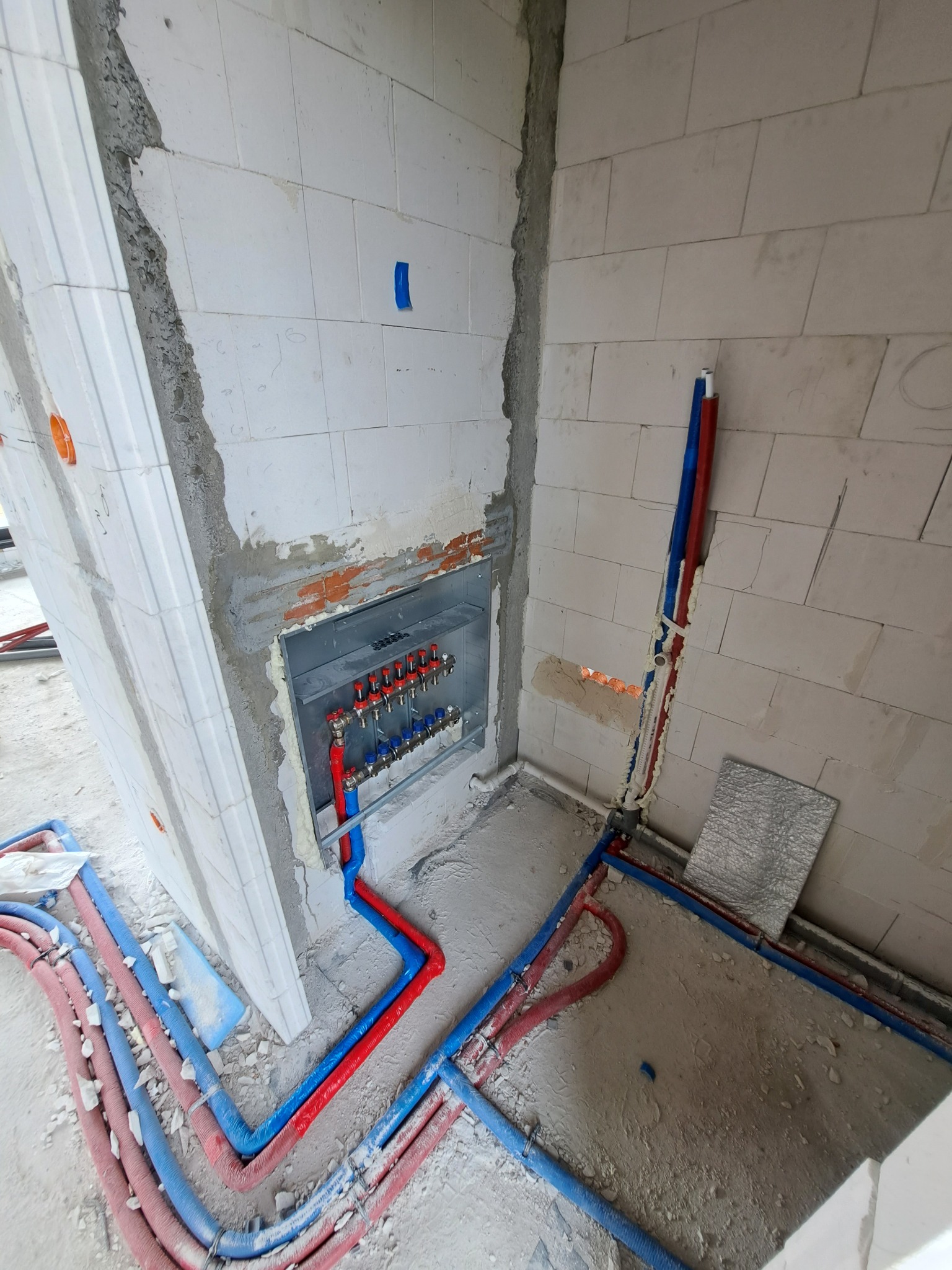Rozdzielacz ogrzewania podłogowego w trakcie montażu w ścianie z betonu komórkowego, widoczne czerwone i niebieskie rury doprowadzające wodę, zabezpieczone pianką montażową.