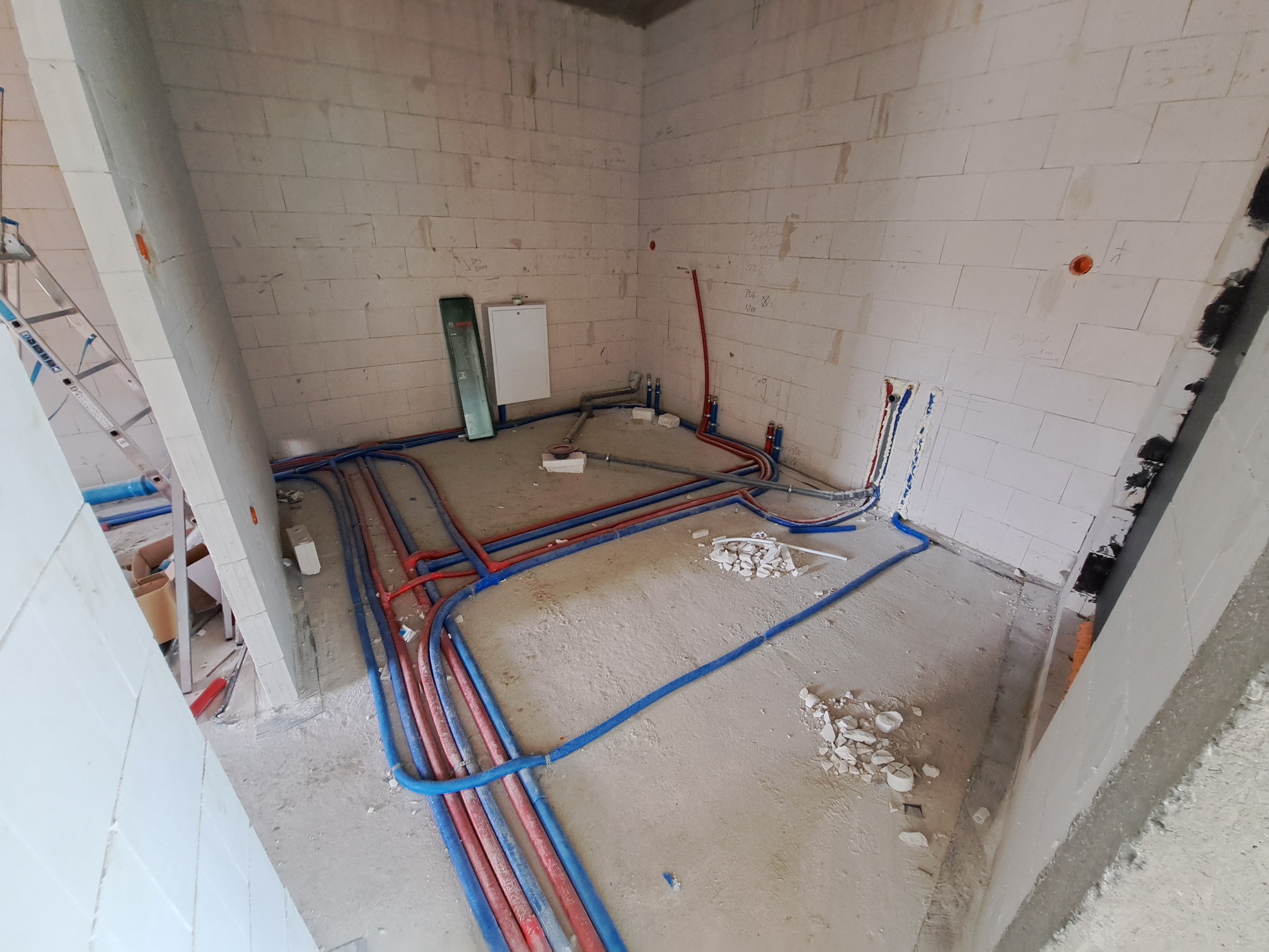 Surowy stan: Instalacja wodna i grzewcza z rur PEX w kolorach czerwonym i niebieskim, ułożona na podłodze w pomieszczeniu z białych bloczków betonowych przed tynkowaniem, widoczna drabina...