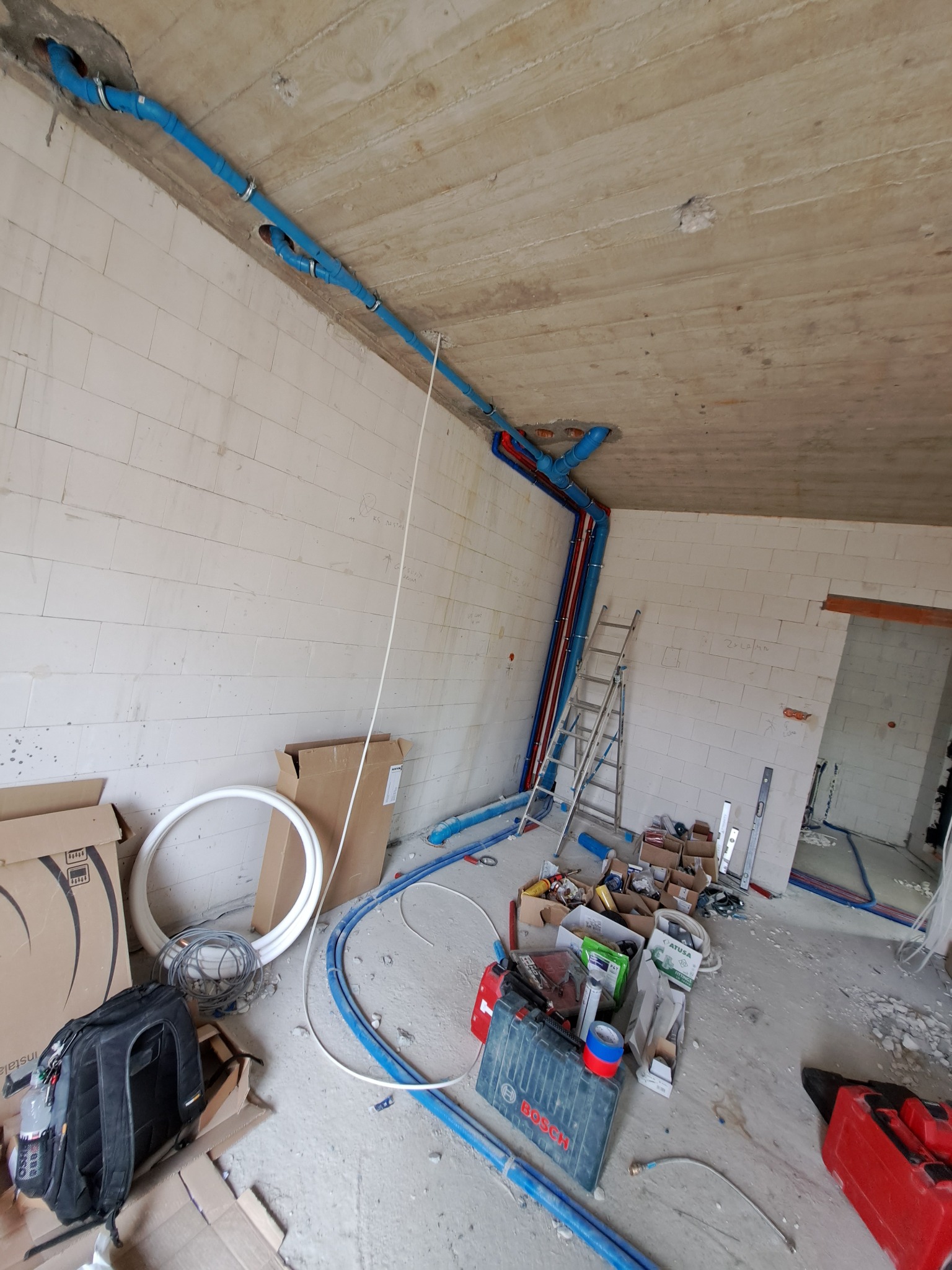 Wnętrze w trakcie instalacji hydraulicznej: niebieskie rury rozprowadzone po suficie i ścianach surowego budynku, drabina oparta o ścianę, narzędzia i materiały budowlane rozłożone na podłodze.