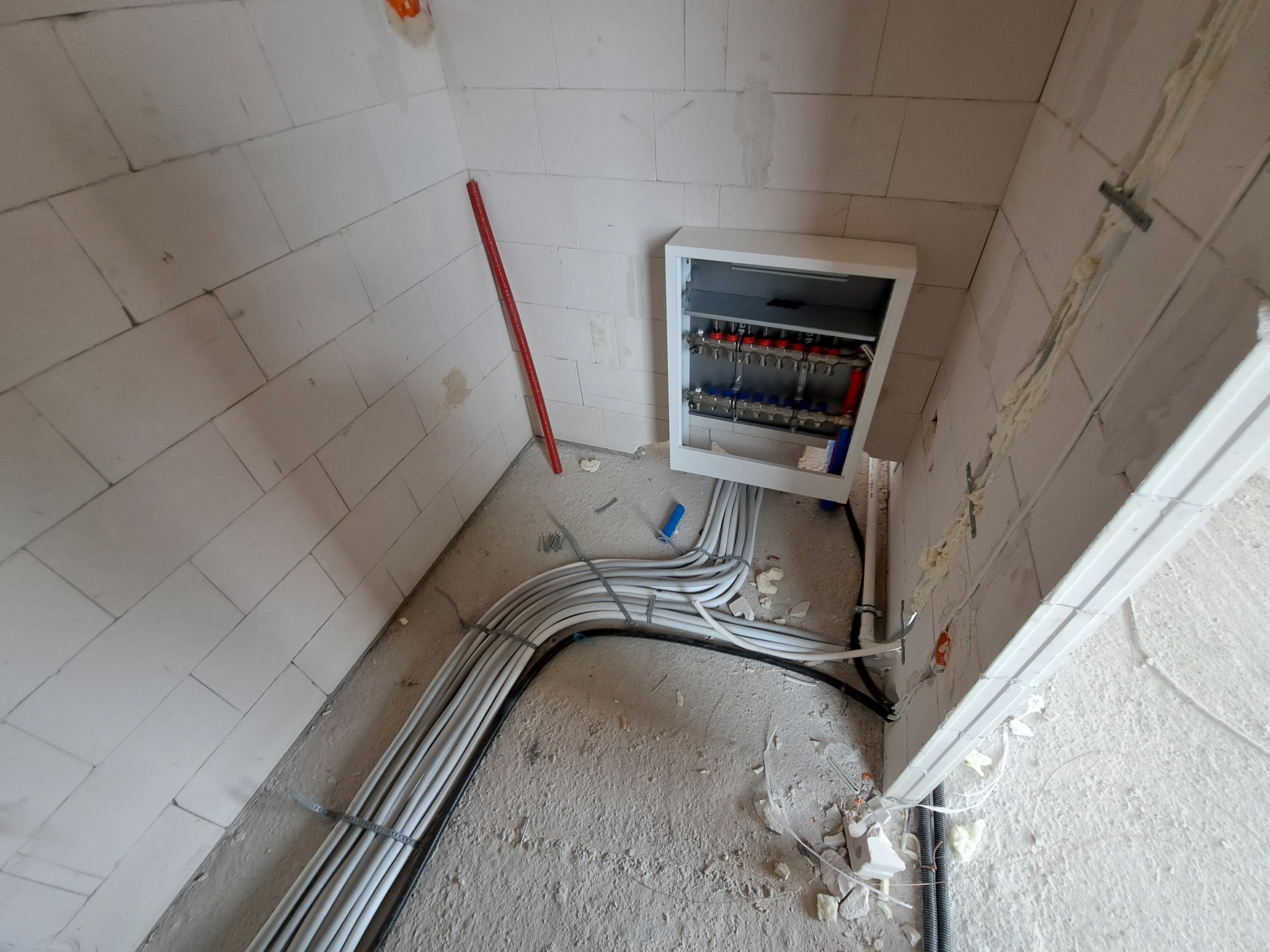 Rozdzielacz instalacji grzewczej w białej obudowie, zamontowany na ścianie z białych bloczków, z poprowadzonymi rurami w peszlach, poziomica oparta o ścianę, surowy stan wykończenia.