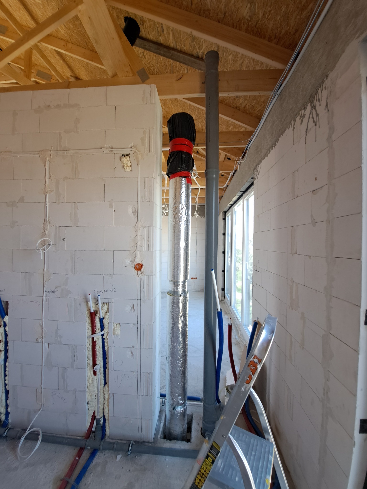 Szara rura wentylacyjna w izolacji aluminiowej, zabezpieczona czarną folią i czerwoną taśmą, przechodzi przez surowy budynek z białych bloczków, widoczne wystające rury instalacji hydraulicznej...