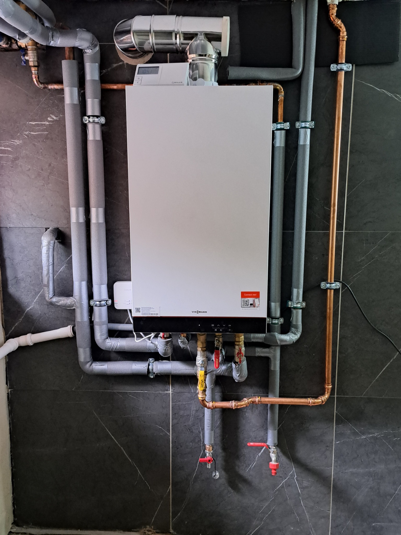 Kocioł grzewczy Viessmann z orurowaniem miedzianym i izolowanym, zamontowany na ścianie wyłożonej ciemnymi płytkami imitującymi marmur.