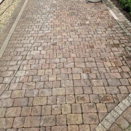 ZAREMBA MYCIE CIŚNIENIOWE Katarzyna Zaremba - Częściowo wyczyszczona kostka brukowa z widocznym urządzeniem do mycia ciśnieniowego i szczotką do zamiatania, ukazująca różnicę w kolorze między czystą a brudną powierzchnią.