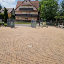 Ułożona, brązowo-żółta kostka brukowa na podjeździe przed domem z otwartą, metalową bramą w słoneczny dzień.