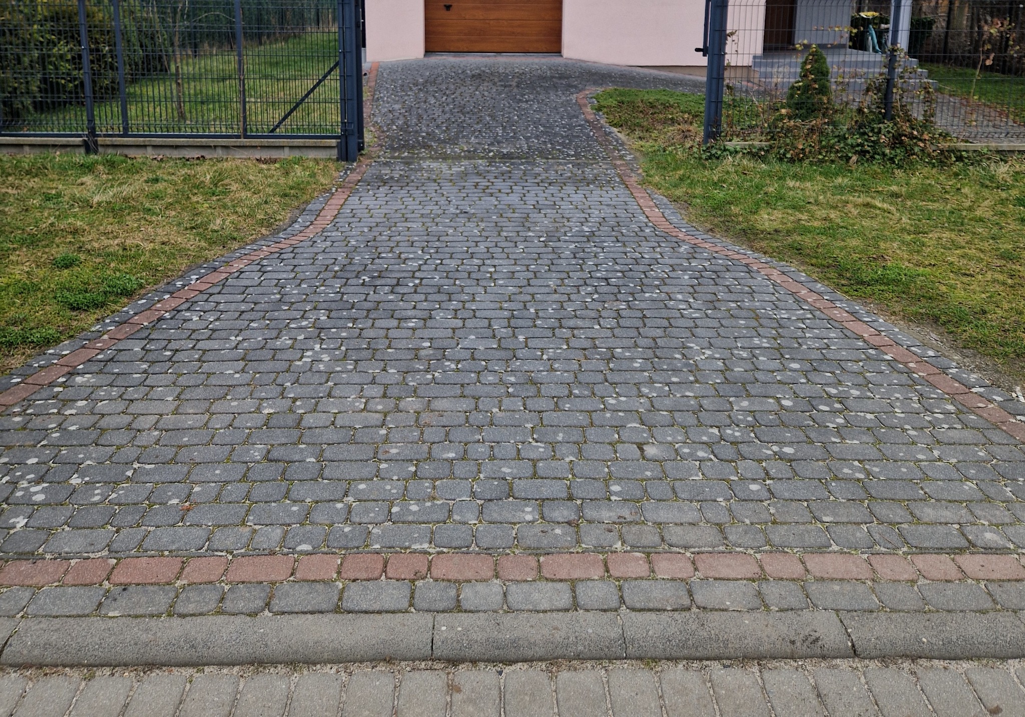 Szeroki podjazd z szarej kostki brukowej z czerwoną obwódką, prowadzący do garażu z drewnianą bramą, widoczny przez bramę w metalowym ogrodzeniu.
