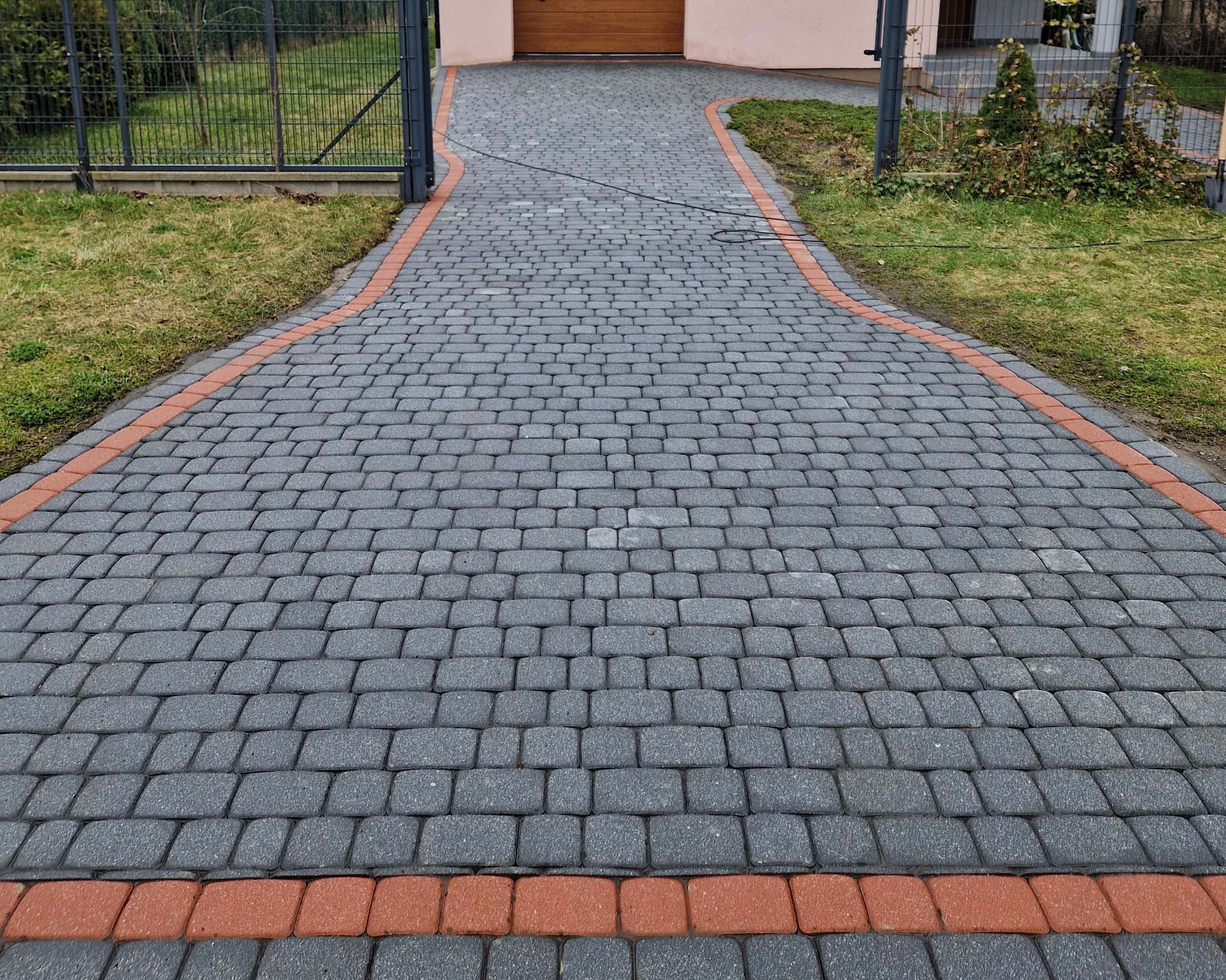 Ścieżka z szarej kostki brukowej z czerwoną obwódką prowadząca do domu z drewnianą bramą garażową, widok z perspektywy wjazdu.