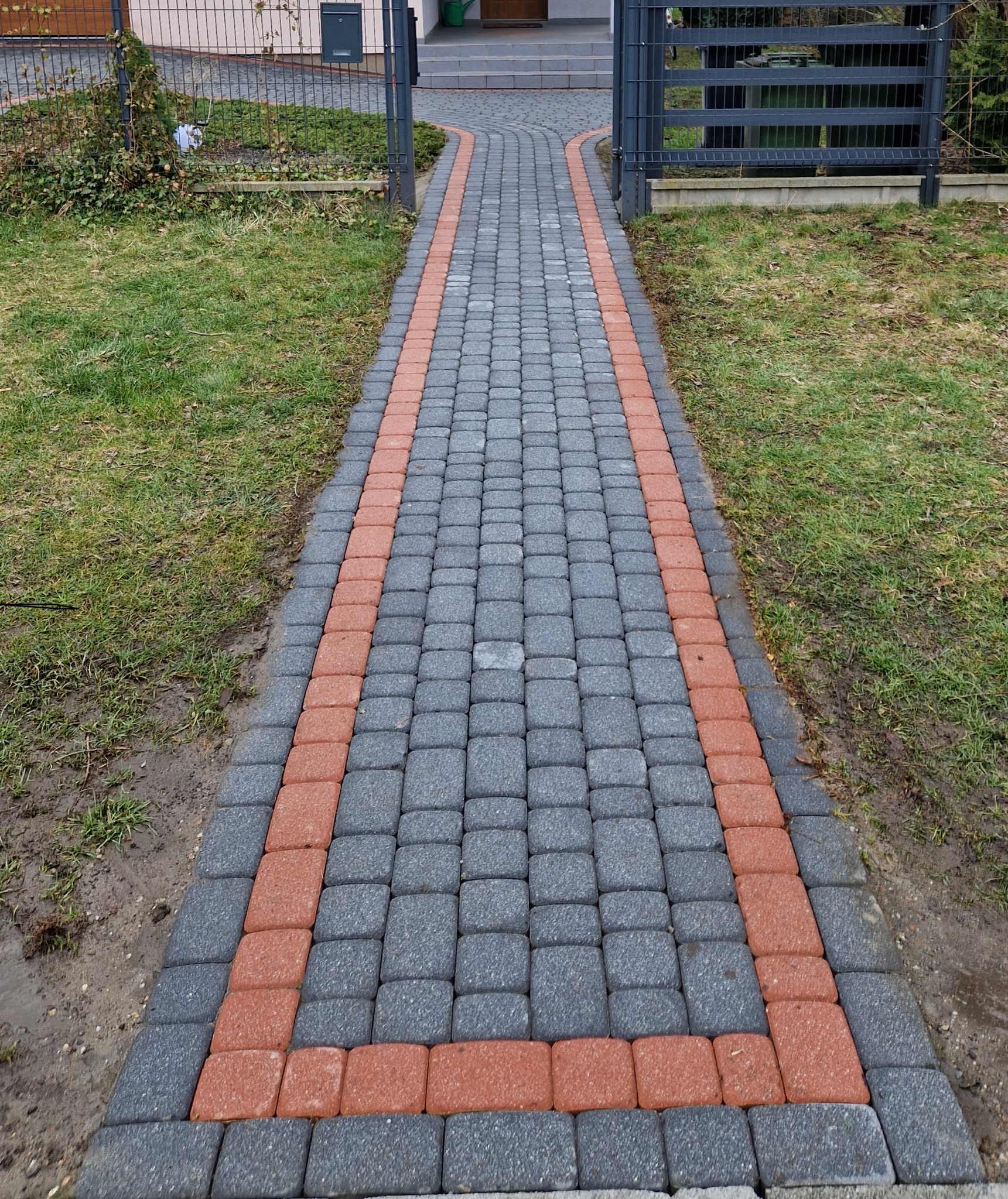 Ścieżka z szarej kostki brukowej z bordowymi obrzeżami prowadząca do furtki i domu, widok z perspektywy osoby stojącej na ścieżce.