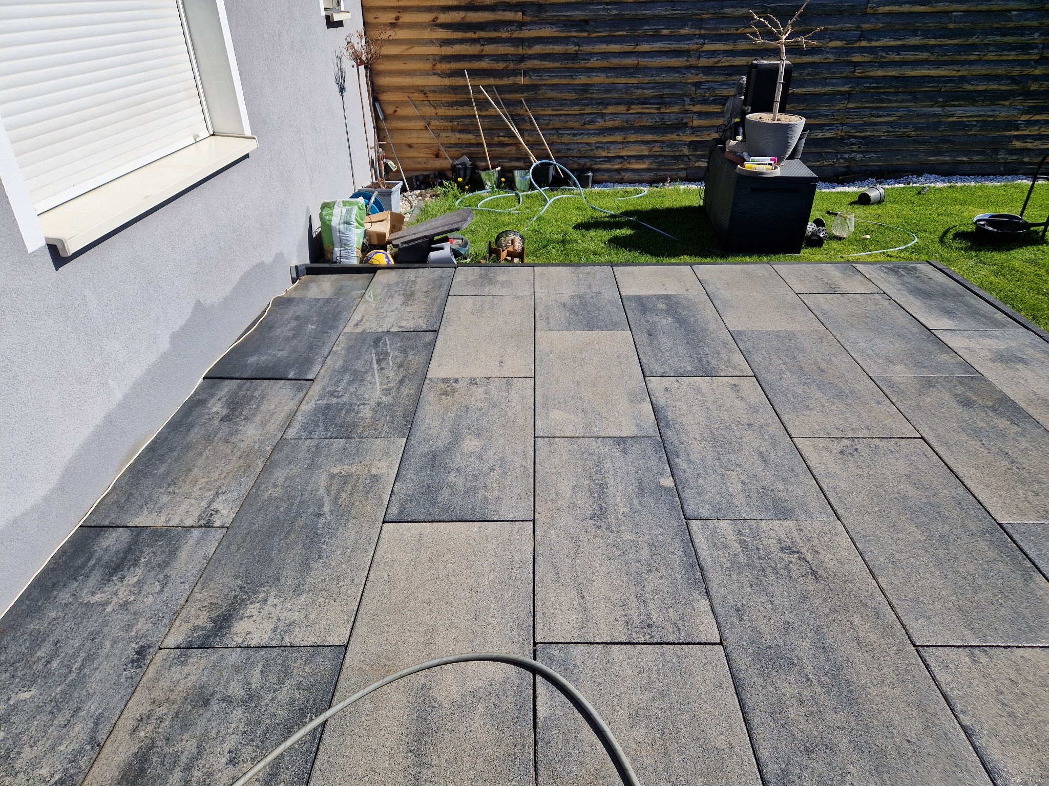 Ułożona kostka brukowa w odcieniach szarości i beżu przylegająca do ściany budynku, widoczne narzędzia i materiały budowlane na trawie w tle.