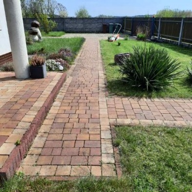 Ułożona z kostki brukowej ścieżka w ogrodzie, z widoczną kolumną domu po lewej i trawnikiem z krzewem po prawej, w tle ogrodzenie i kosiarka.