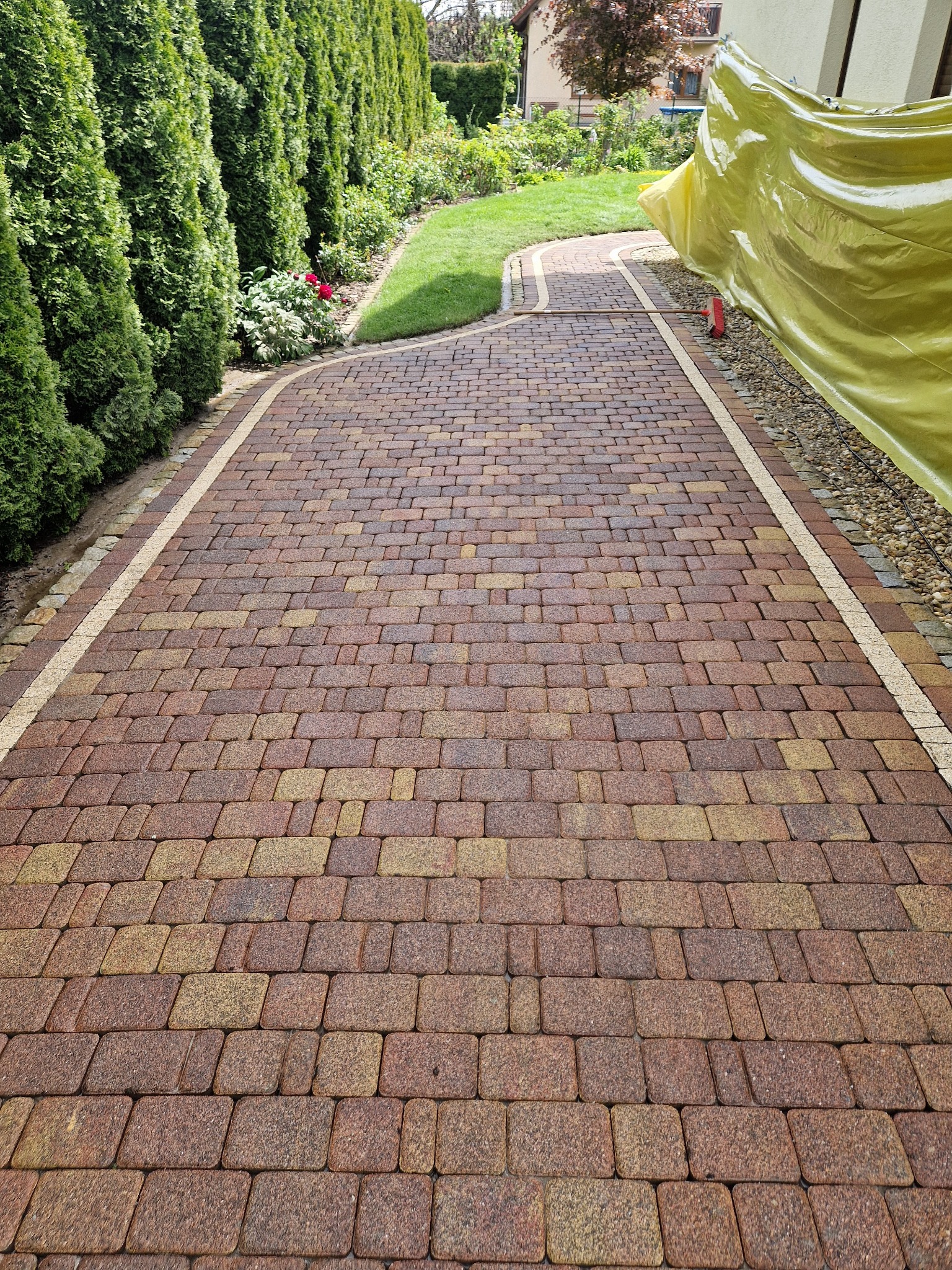 Ułożona z kostki brukowej ścieżka ogrodowa w odcieniach brązu i beżu, obramowana jasnymi paskami i zielenią, z widoczną folią ochronną na budynku w tle.