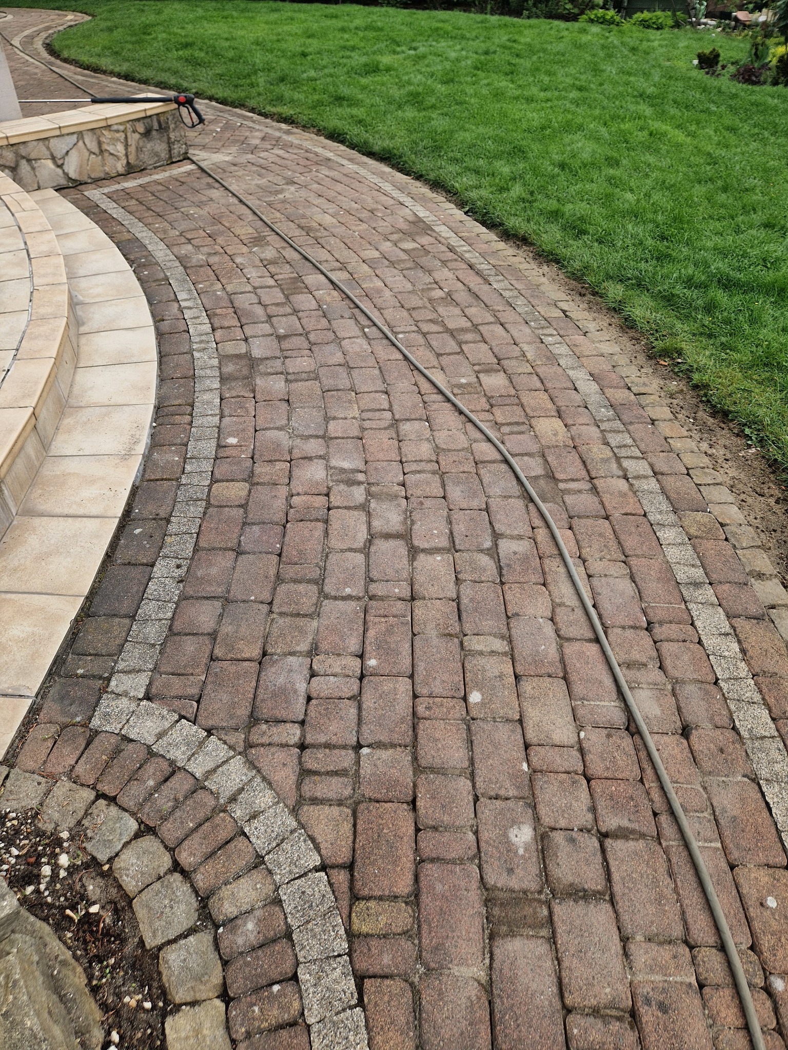 Zakrzywiona ścieżka z czerwonej kostki brukowej z szarym obramowaniem, częściowo umyta, z wężem ciśnieniowym leżącym na powierzchni, prowadząca do zielonego trawnika.