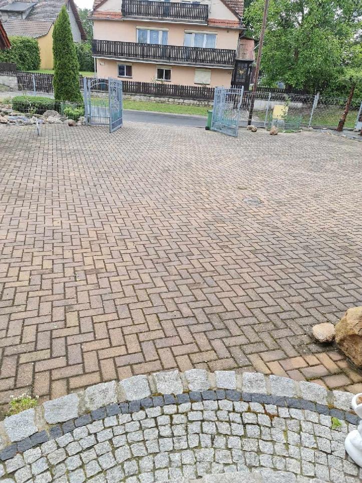 Szeroki kadr na kostkę brukową ułożoną we wzór jodełki, z widocznymi śladami zabrudzeń, z elementami dekoracyjnymi z kamienia na pierwszym planie i otwartą bramą w tle.