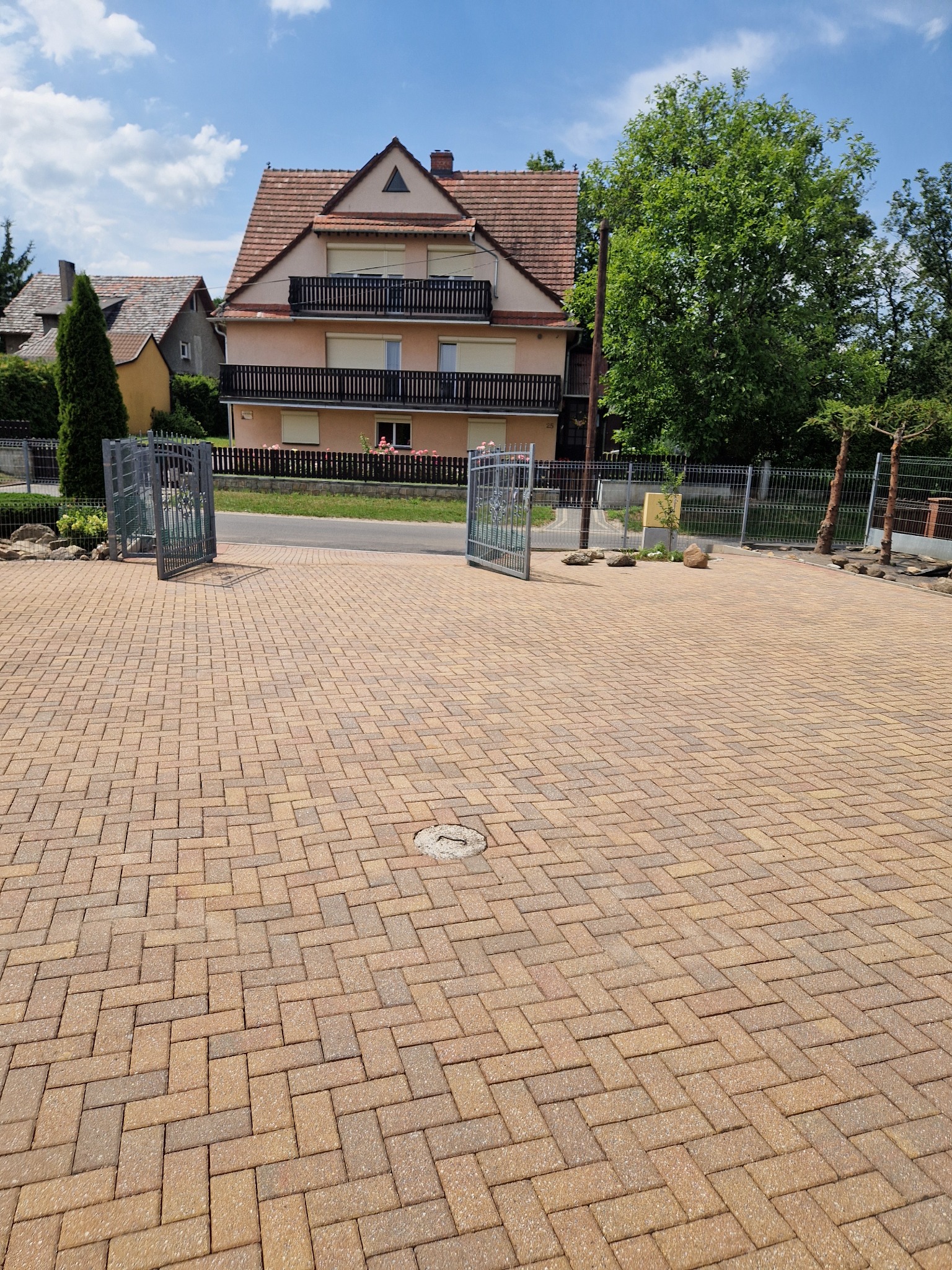 Ułożona, brązowo-żółta kostka brukowa na podjeździe przed domem z otwartą, metalową bramą w słoneczny dzień.